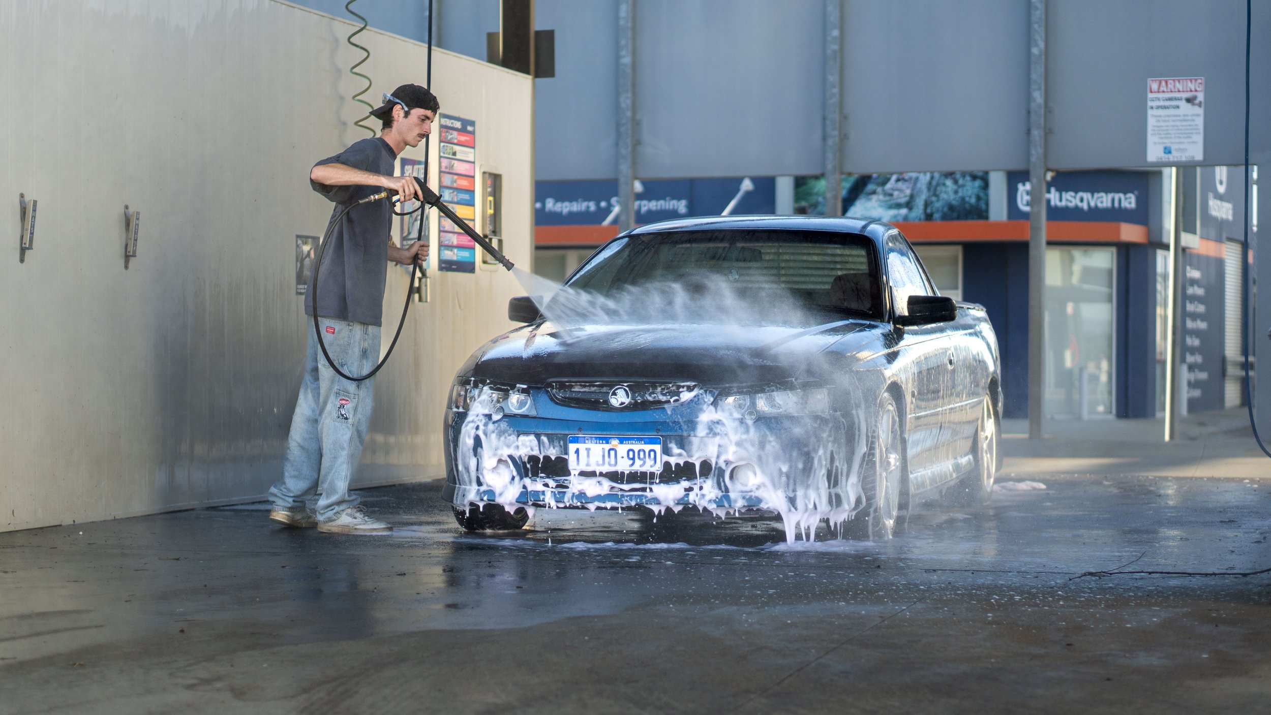 A young man wearing a black cap and grey t-shirt is washing a black car with a hose, creating soap suds on the front of the vehicle. The scene is at a car wash station with a blue and grey background.