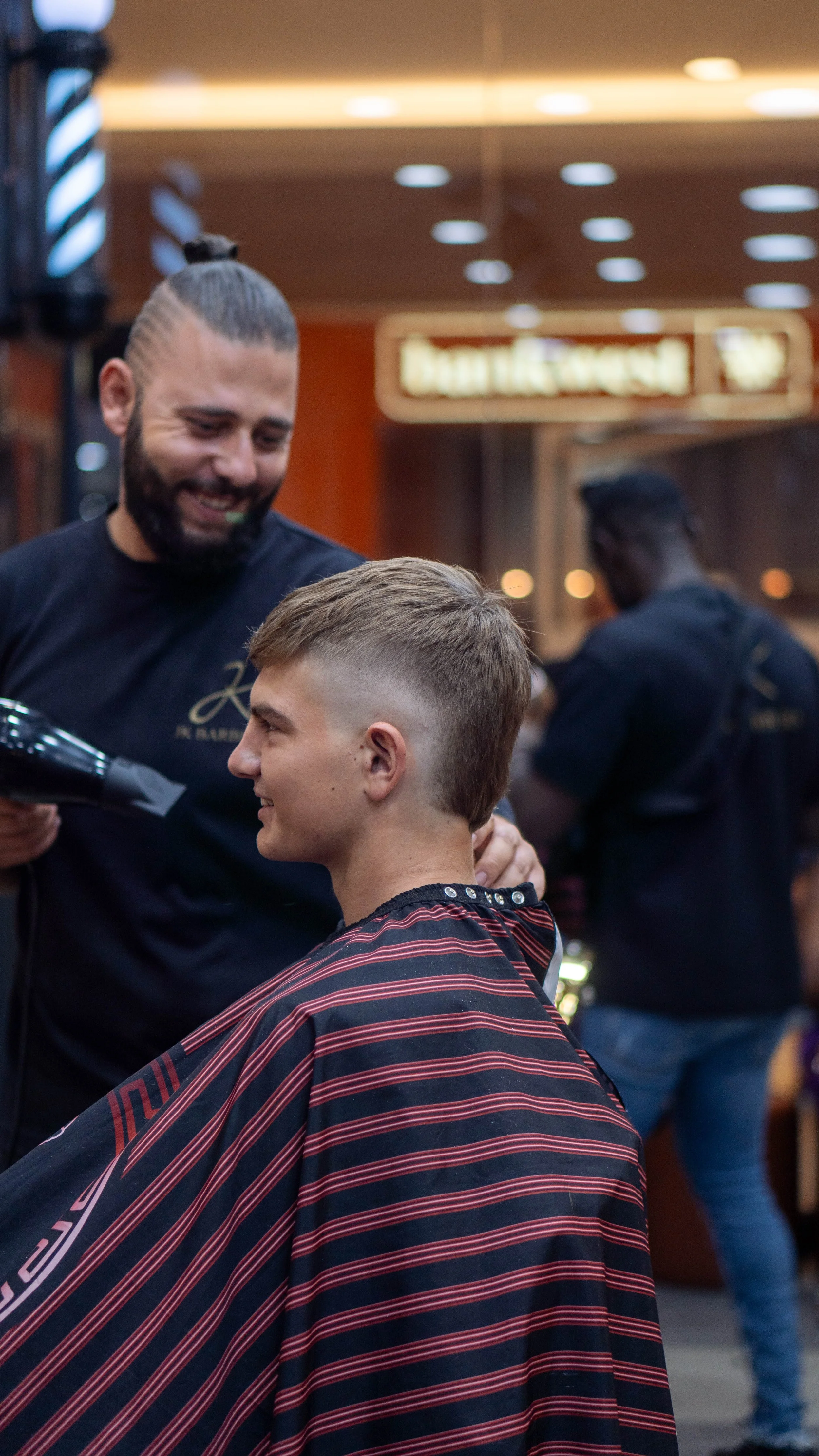 Barber smiling while using a hairdryer on a young man's hair in a barbershop.