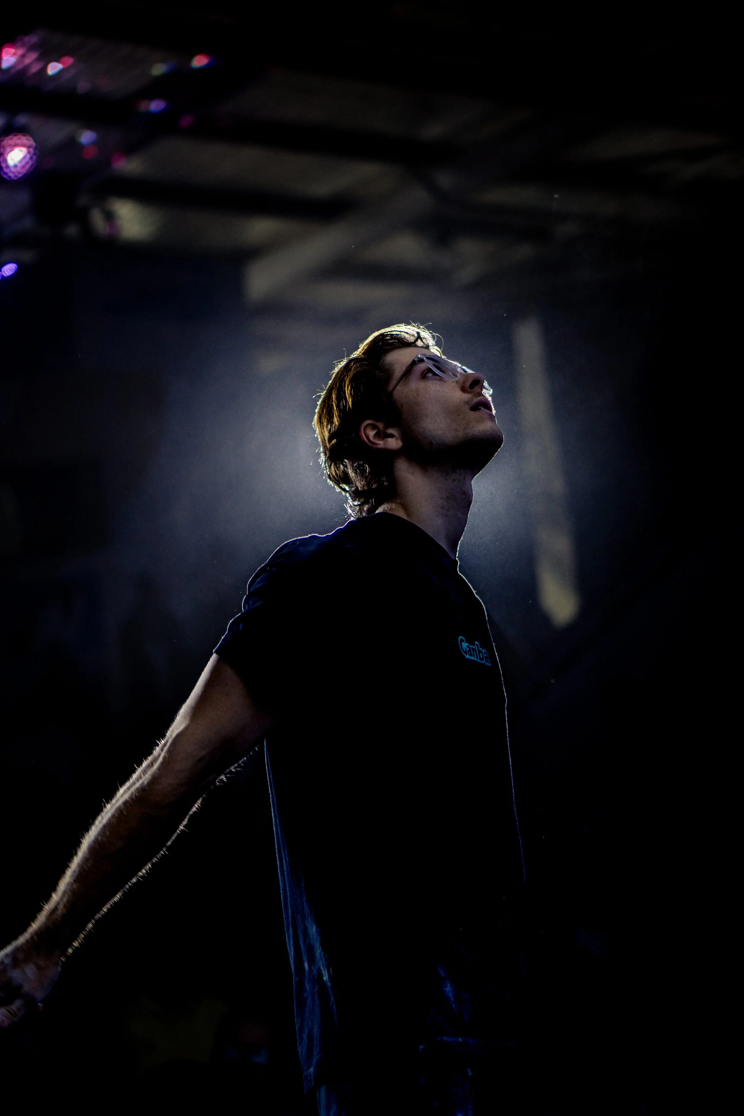 Young man with glasses, facing upwards, illuminated by a spotlight, with water streams splashing around him at a waterpark or similar venue.