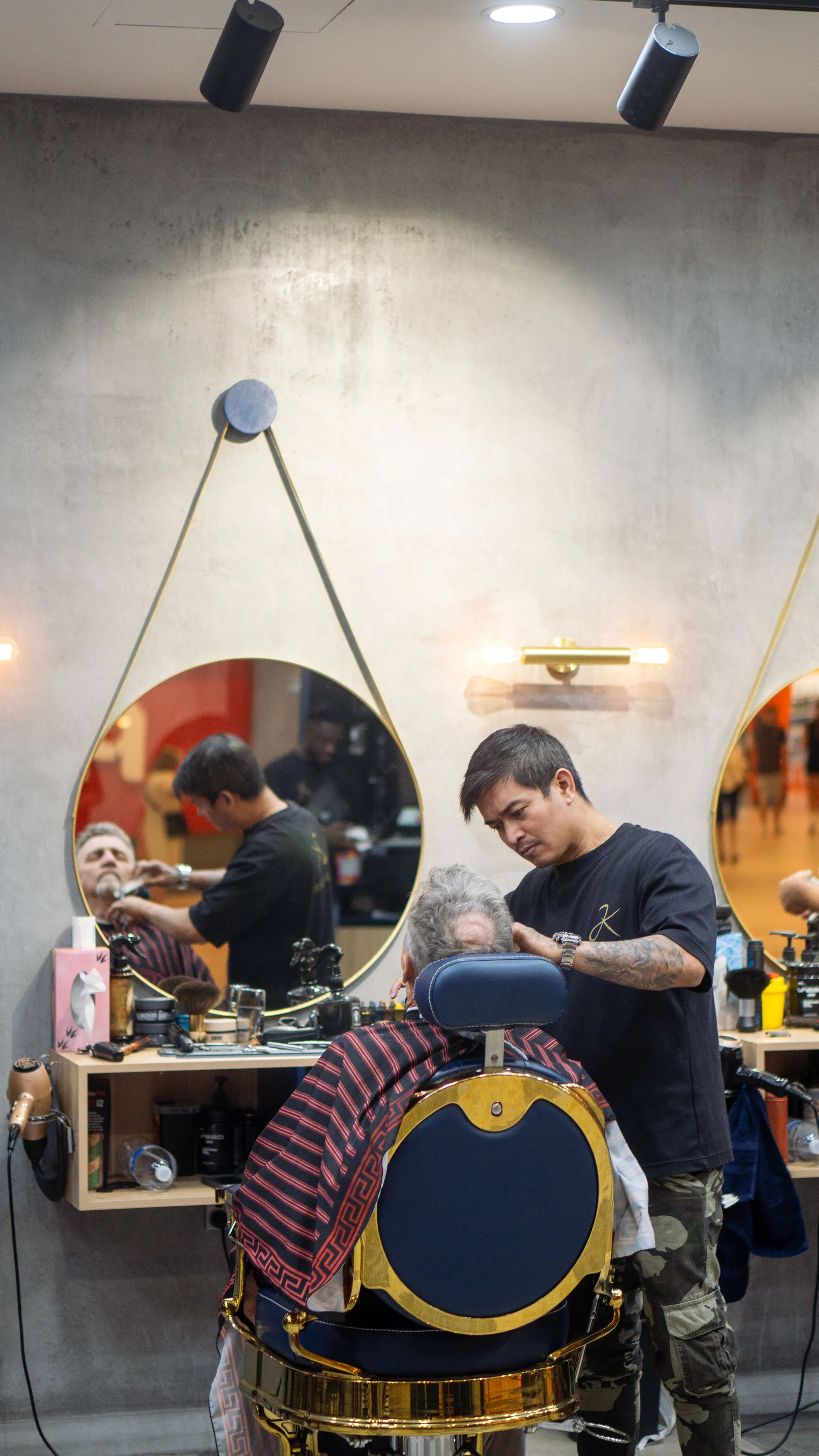 A barber is cutting a man's hair in front of a large mirror with two round, gold-framed mirrors on a textured wall. The barber's station has various grooming tools, and there is a barber's chair with a patterned cape on the client.