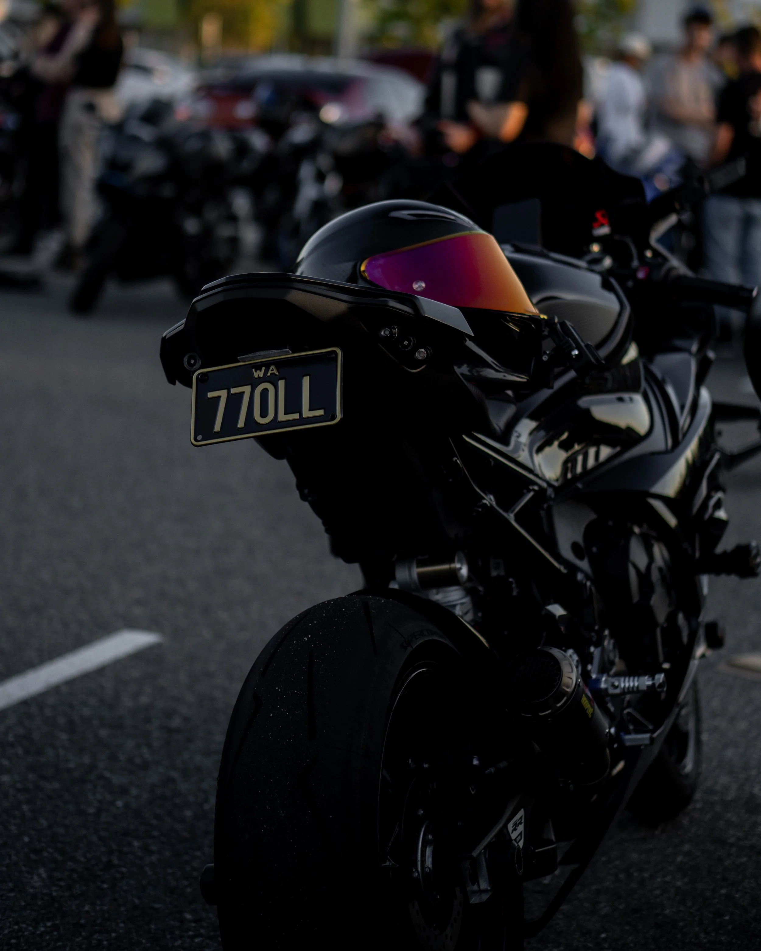 Black motorcycle with a tinted helmet on the seat, parked on a road with blurred people and cars in the background during evening or dusk.