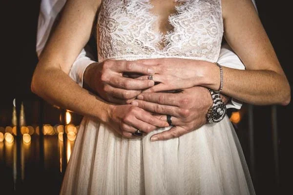 Close-up of a newlywed couple embracing with their wedding rings visible against warm evening lights.
