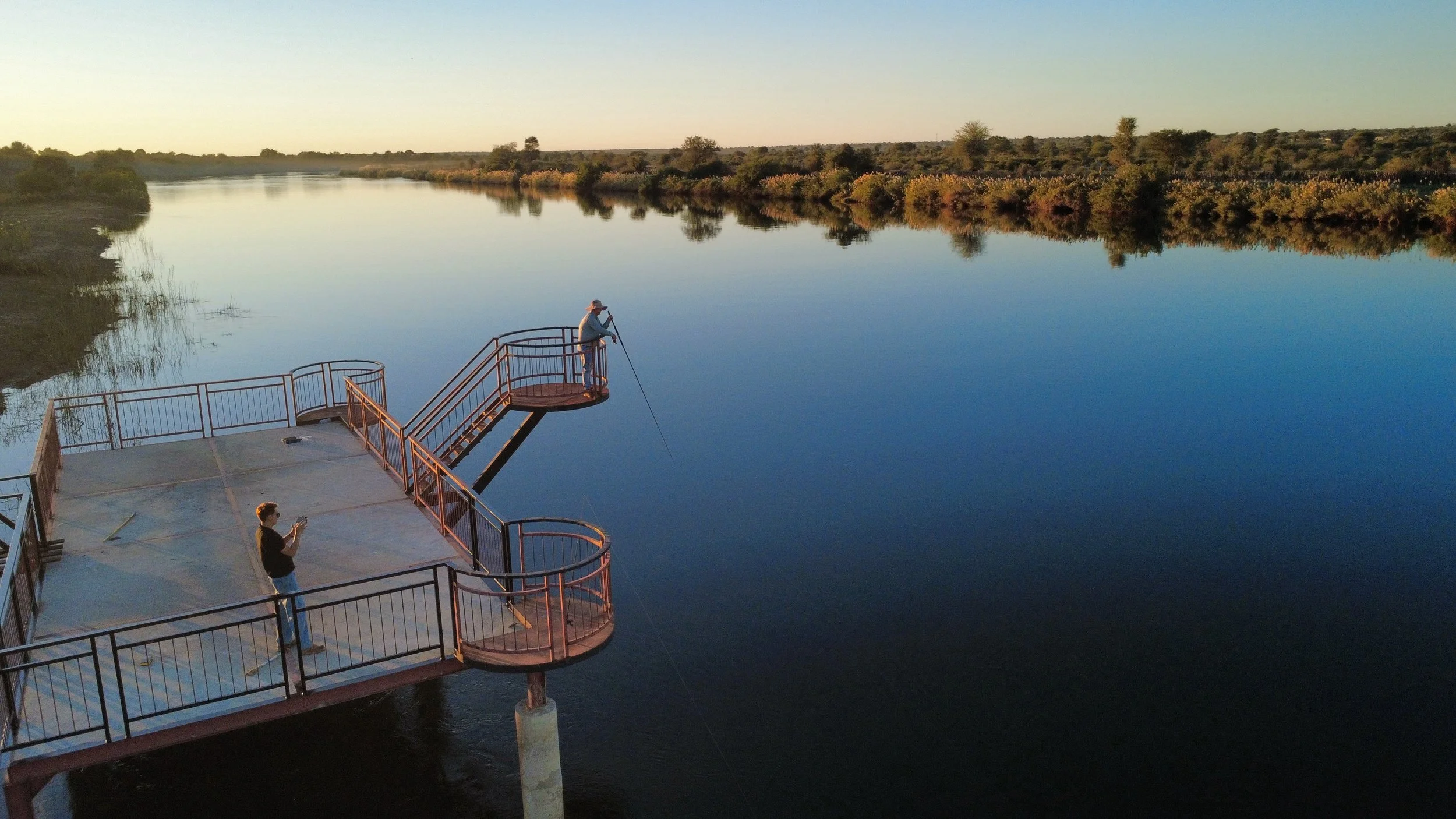 People fishing off a pier on a calm river during sunset with trees in the background.