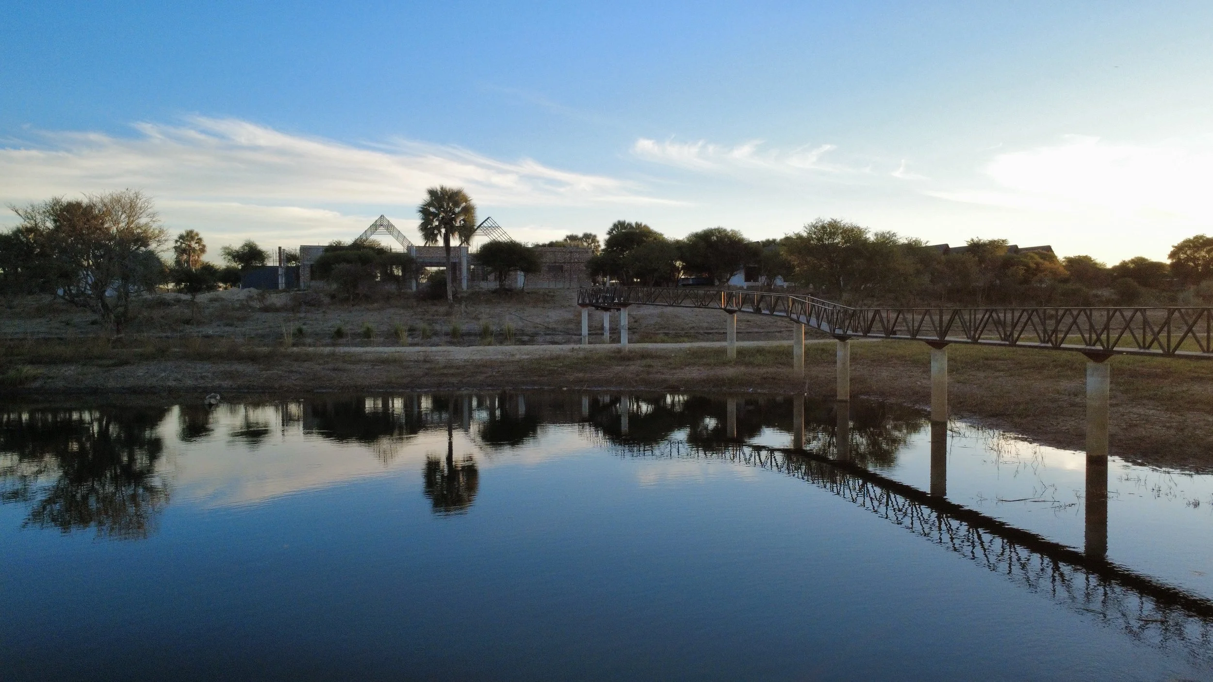 A peaceful outdoor scene features a calm body of water reflecting the sky, trees, and a bridge. In the background, there are some buildings under construction or renovation and a clear sky with a few clouds.