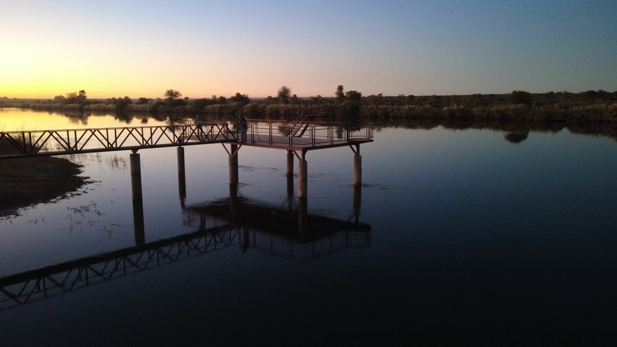 A scenic view of a lake at sunset with calm water reflecting the sky. A wooden walkway extends over the water with a few people standing on it. The background features sparse trees and a clear sky transitioning from yellow to blue.