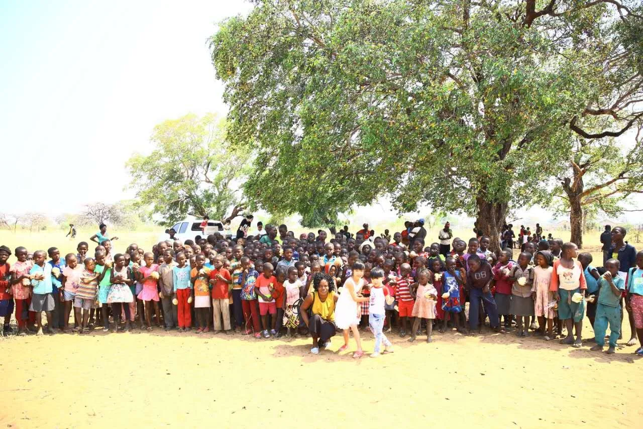 A large group of children and adults gathered outdoors under a big tree in a rural area, with open fields and a few smaller trees in the background.