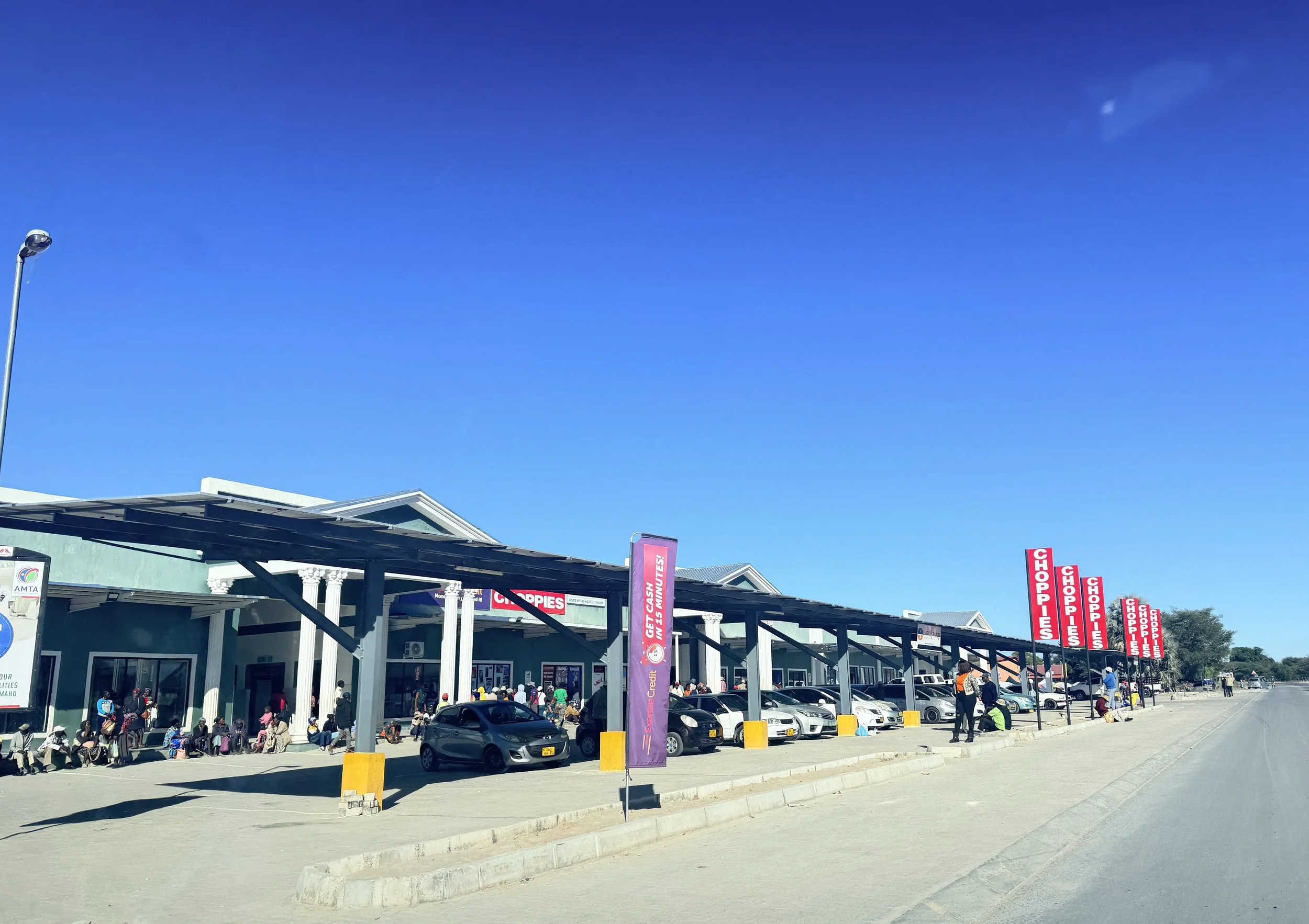 Shopping center with parking lot and people waiting in line, discount signs for chips and chocolates displayed on red banners, clear blue sky, some cars parked and a few individuals walking.