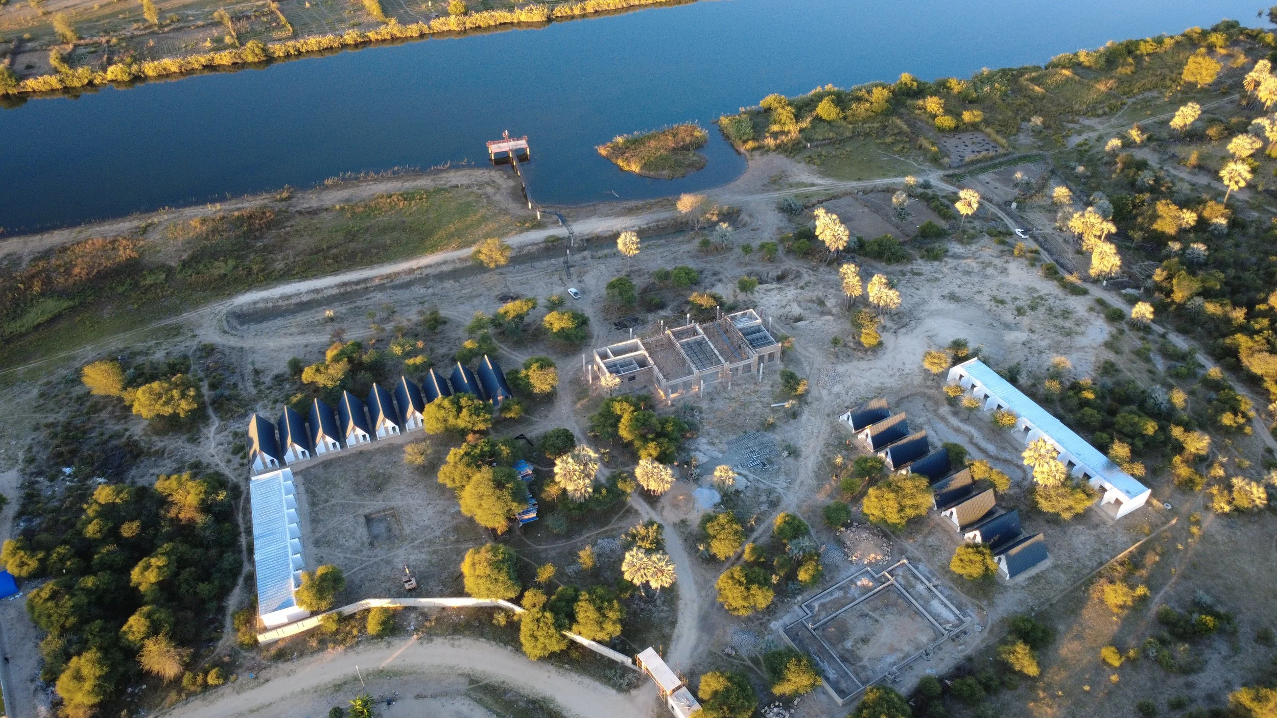 Aerial view of a lakeside area with a dock, trees, pathways, and several buildings under construction or renovation.
