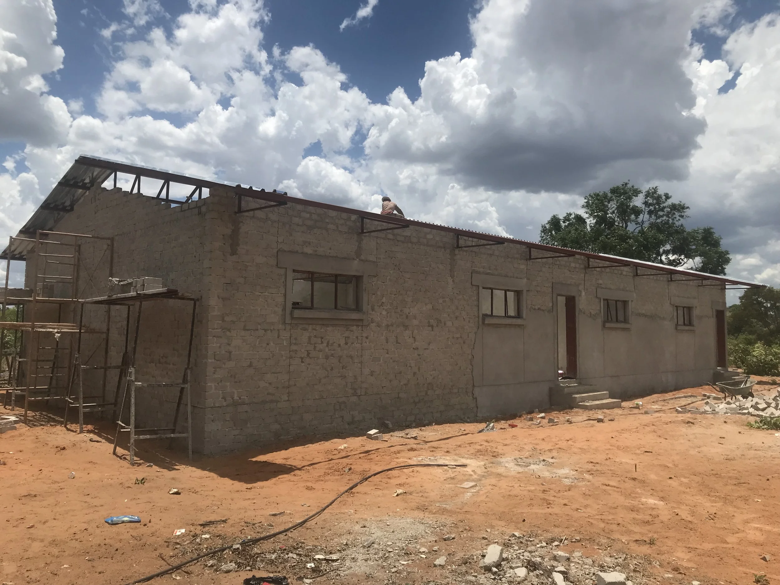 A building under construction with a brick exterior and windows, scaffolding on the left side, and a worker on the roof, the classroom under construction