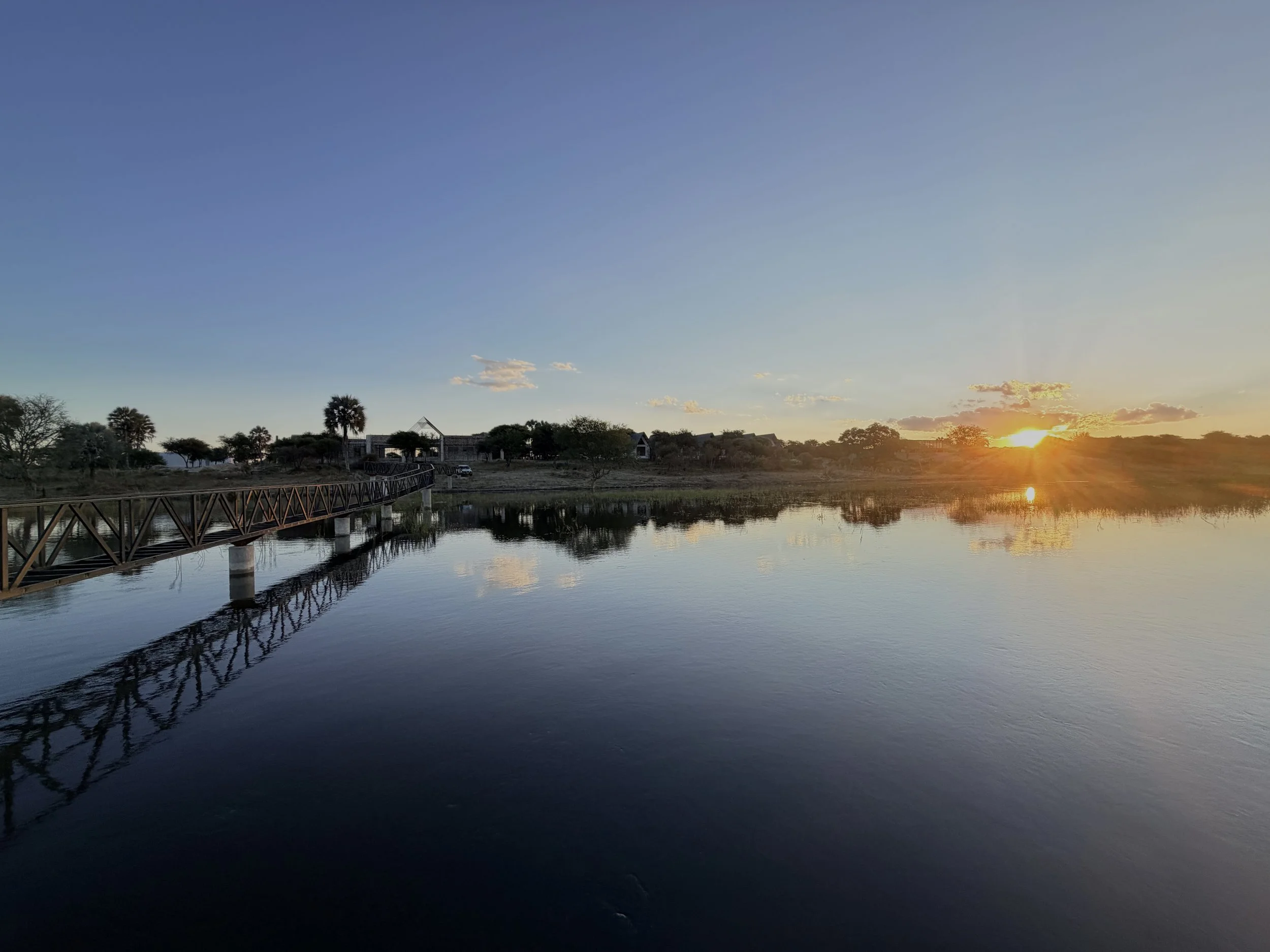 Sunset over a calm body of water with trees and buildings in the background, and a wooden bridge reflected on the water.