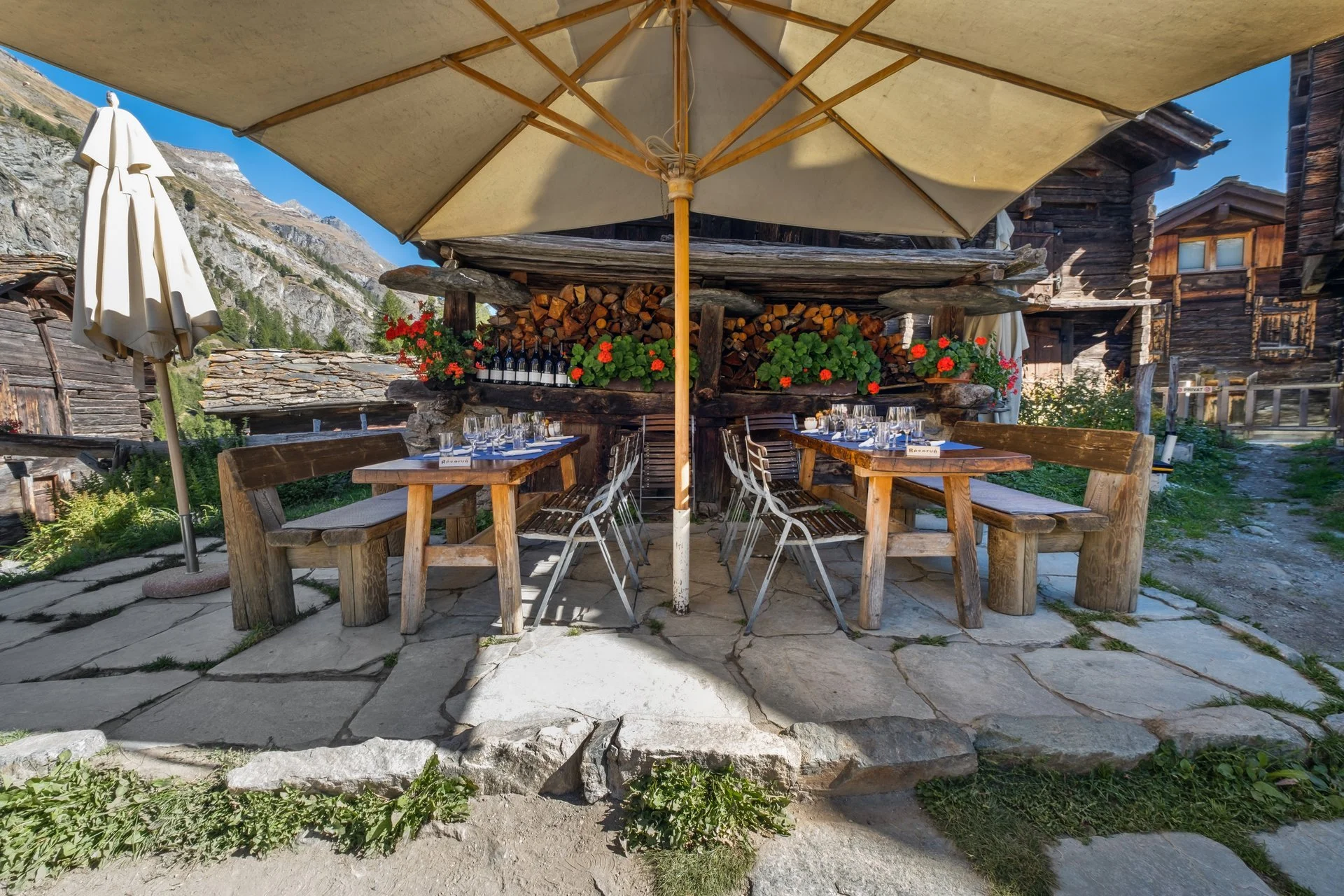 Ein rustikaler Outdoor-Tisch im Schatten eines Sonnenschirms vor einer holzverkleideten Berghütte, mit Weingläsern und Blumen, in den Bergen bei schönem Wetter.