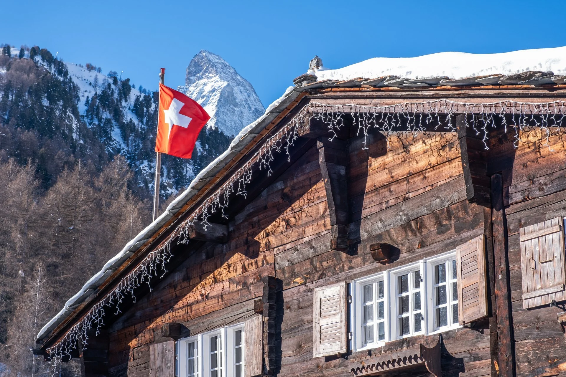 Historisches Holzhaus mit Schweizer Flagge und Weihnachtslichtern, im Hintergrund schneebedeckte Berge und blauer Himmel.