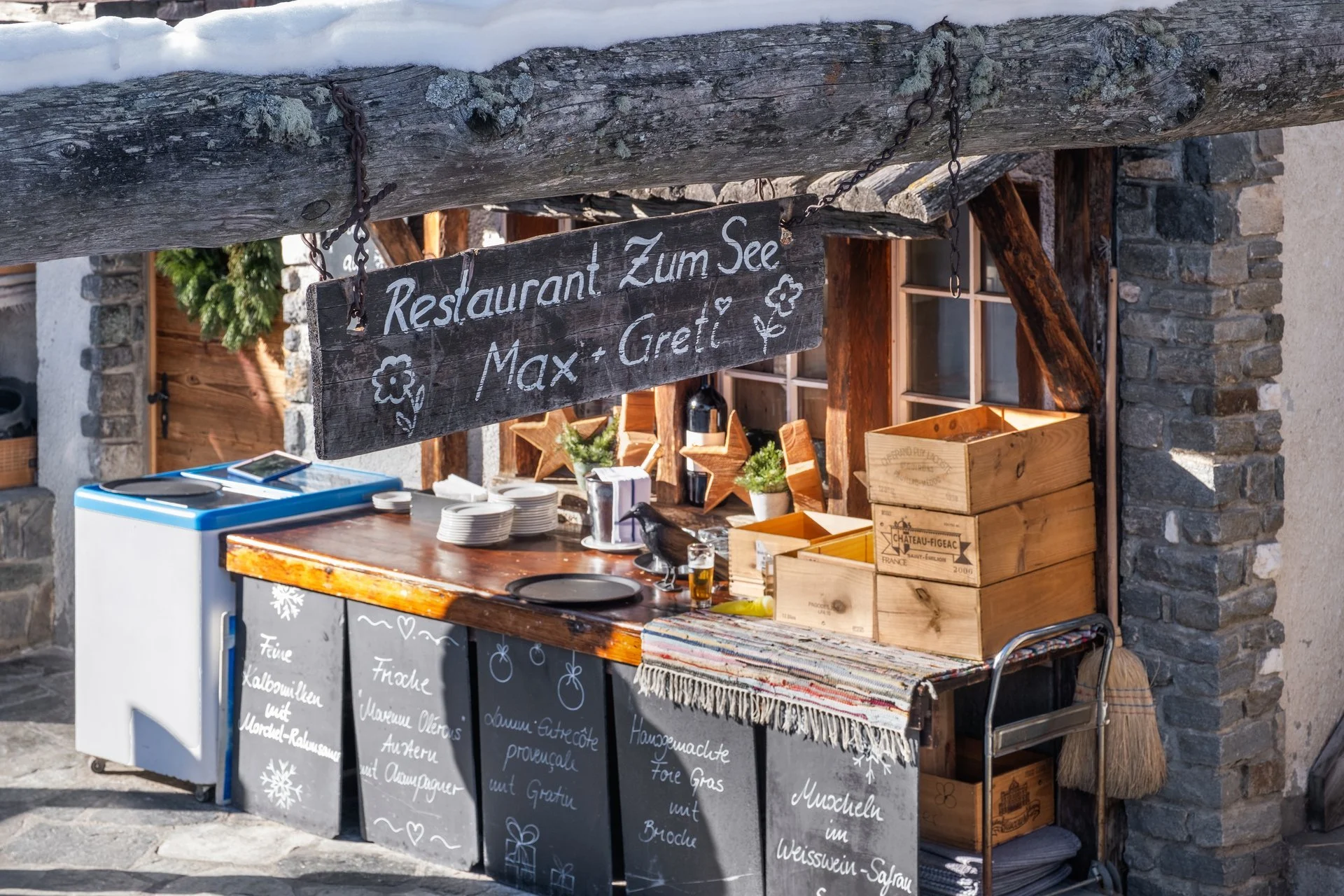 Ein Holzschild mit der Aufschrift ‚Restaurant Zum See Max + Gredi‘ hängt an einem Holzstoß bei einem kleinen Verkaufsstand mit Gläsern, Pflanzen und Holzboxen, vor einer Steinmauer, im Winter mit Schneeregen auf dem Dach.