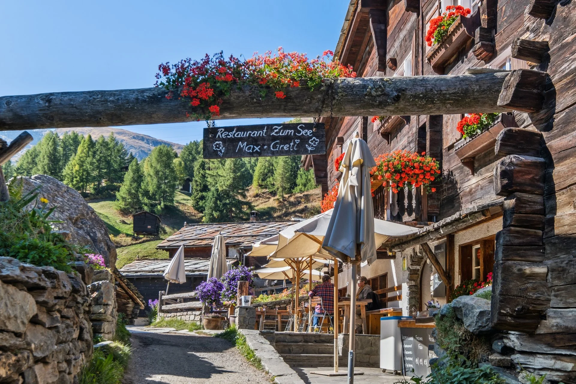 Blick auf ein Restaurant im alpinen Stil mit Holzfassaden, blühenden Blumen und Tischen im Außenbereich unter Sonnenschirmen, umgeben von einer Berglandschaft.