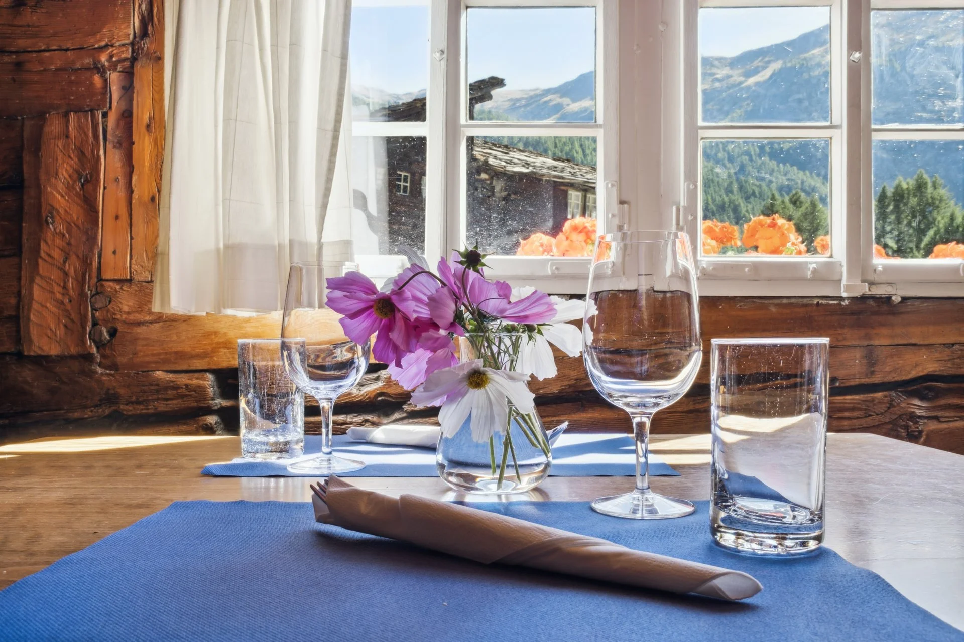 Ein gedeckter Tisch im rustikalen Chalet mit Wasser, Weingläsern und einem Blumenstrauß vor einer Fensteransicht auf Berge und Bauernhäuser.