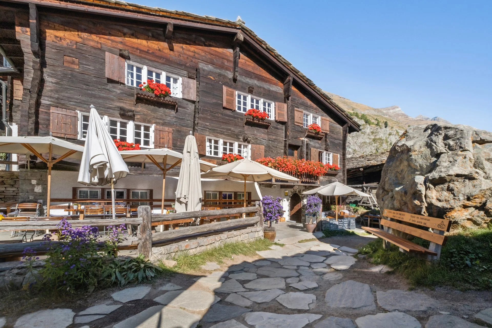 Ein altes, dunkles Holzhaus mit Balkon blumen und Fensterläden, umgeben von Berglandschaft und einem Garten mit Tischen und Sonnenschirmen.
