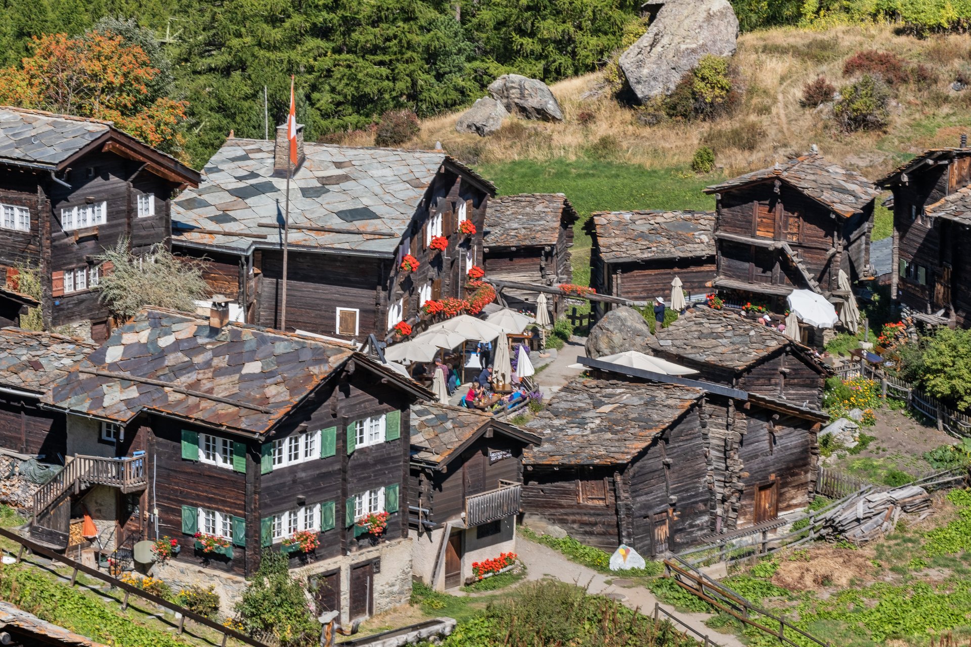 Blick auf traditionelle Holzhäuser in einer alpinen Bergregion mit vielen Menschen, die im Freien sitzen und das schöne Wetter genießen, umgeben von grünen Wäldern und Felsen.
