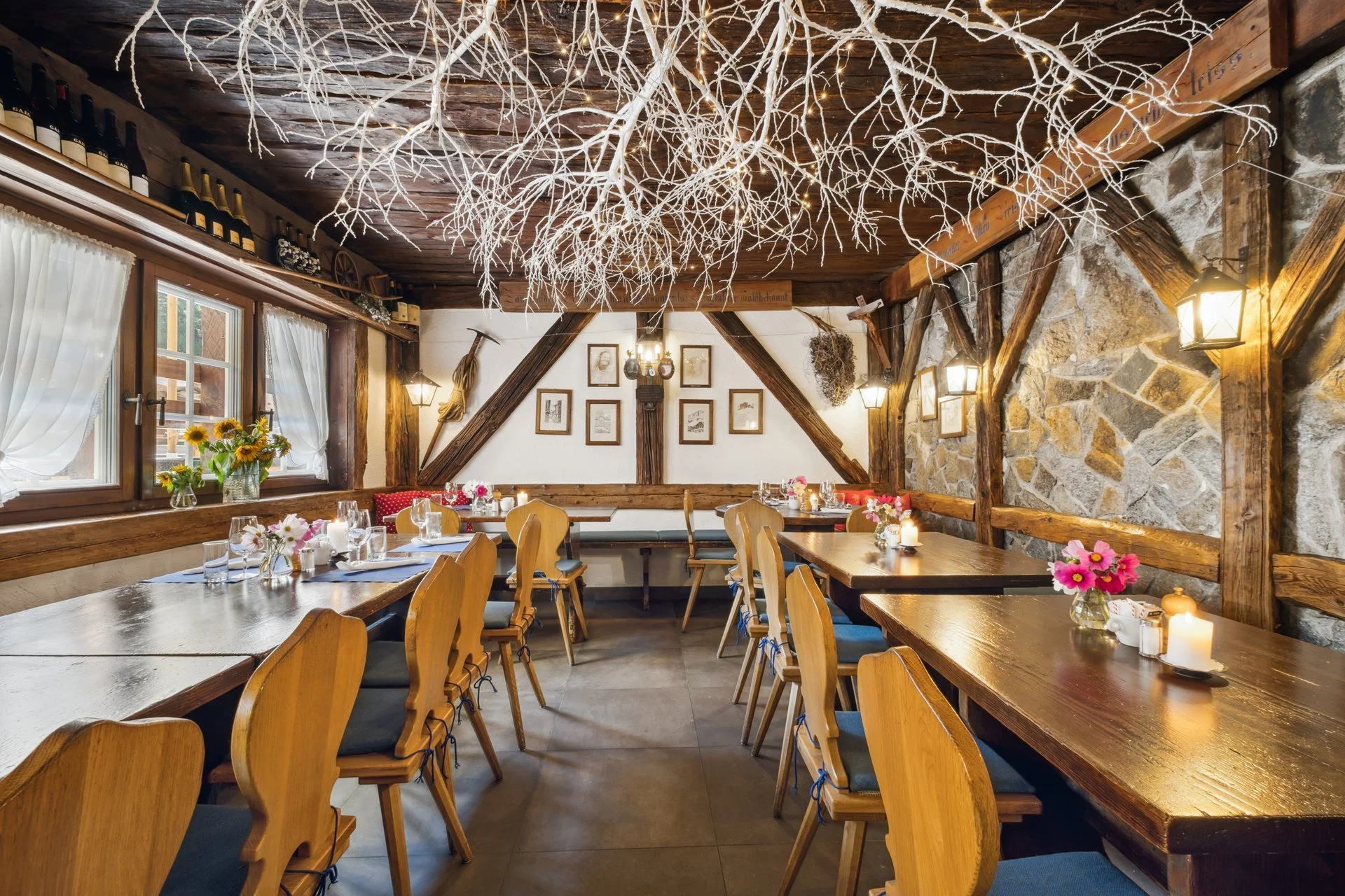 Ein gemütliches rustikales Restaurant mit Holztischen und Stühlen, Fenster mit weißen Vorhängen, Wanddekorationen, einem Kamin und einem Kronleuchter aus weißen Zweigen. Es sind Kerzen und Blumen auf den Tischen dekoriert.