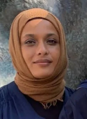 A woman wearing a tan headscarf and a dark blue top, standing outdoors with a rocky background.
