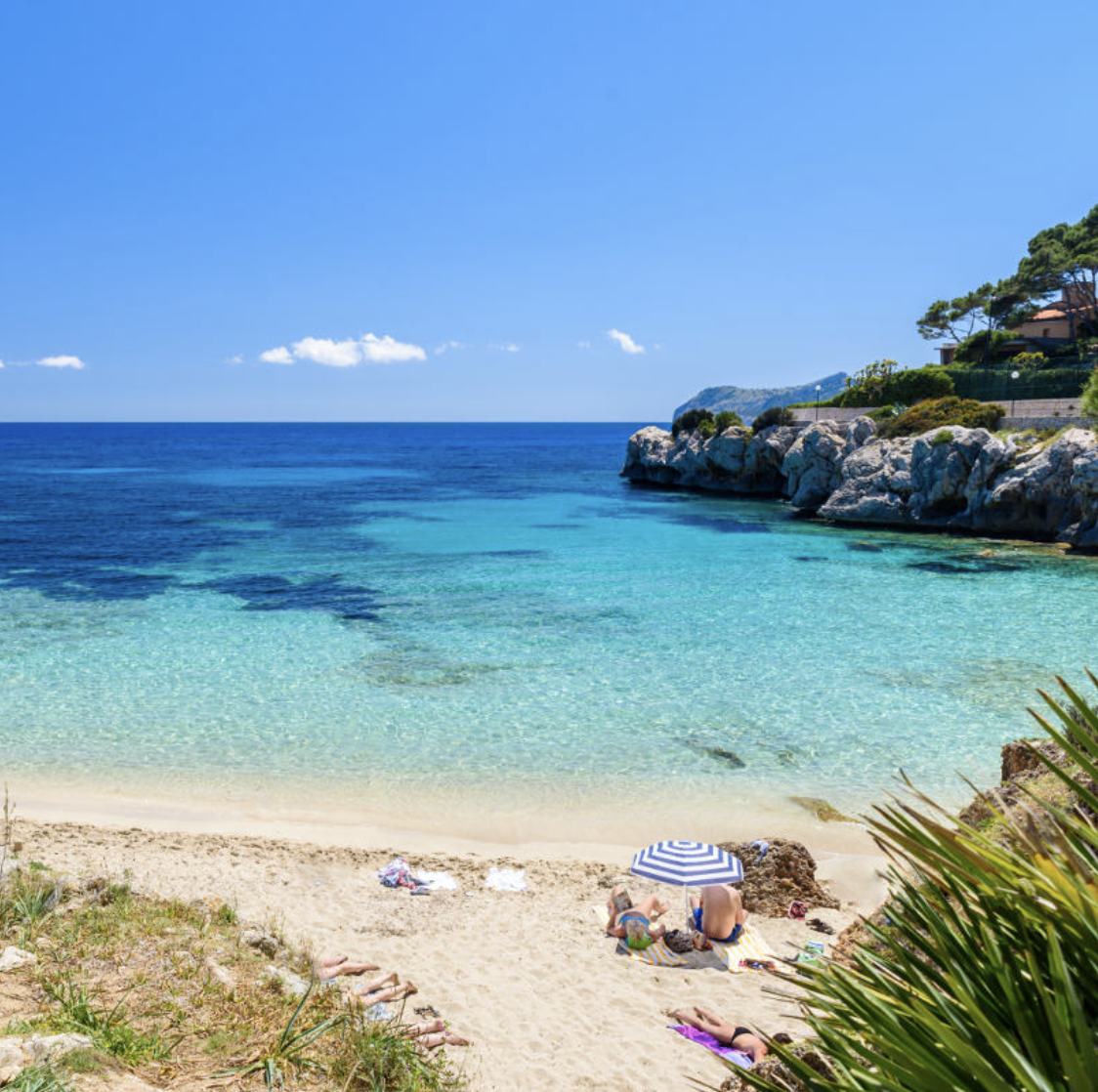 Beach with turquoise water, white sand, rocky coastline, and a couple under a striped umbrella.