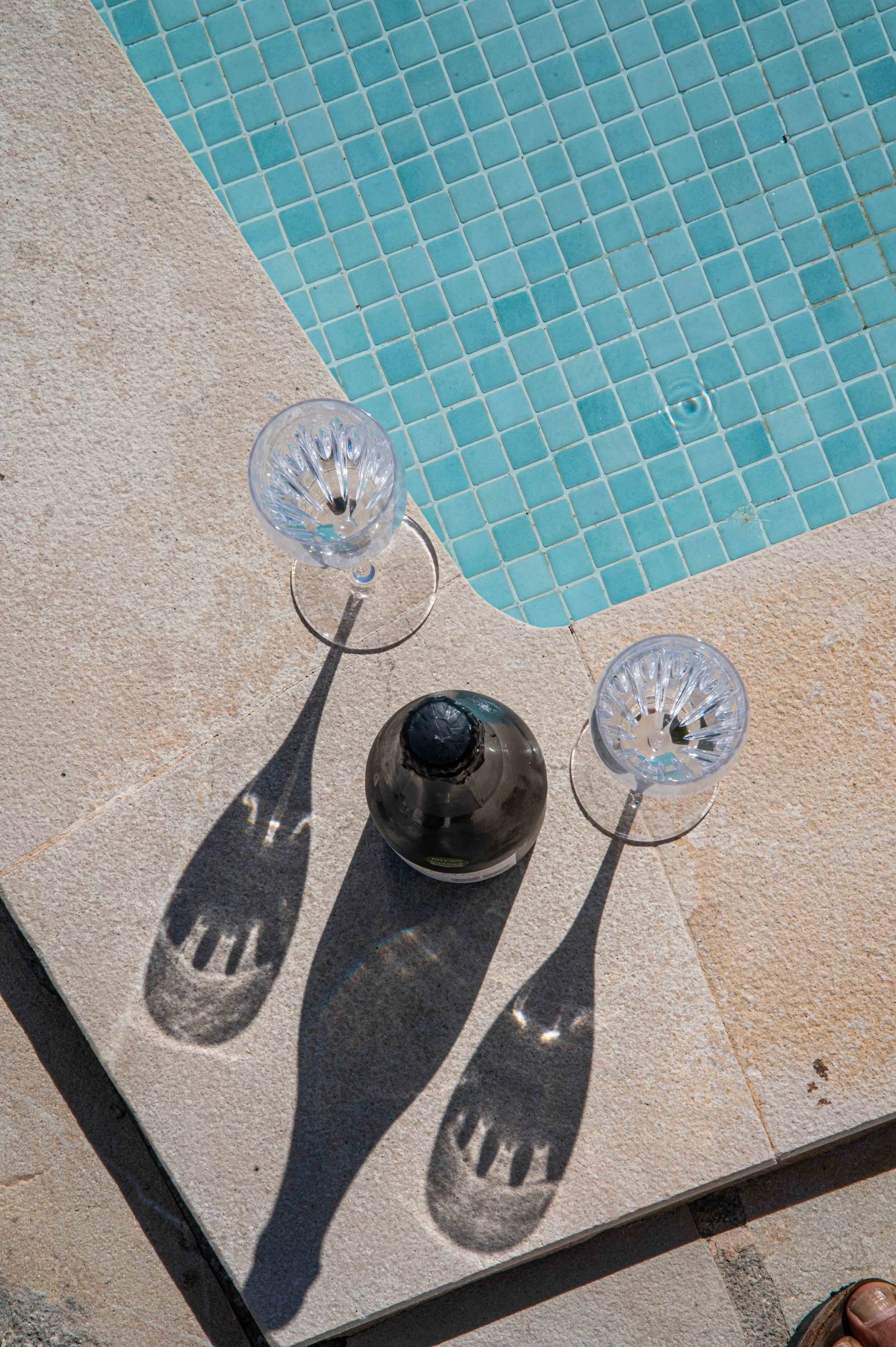 Two empty crystal wine glasses and a wine bottle are placed on a concrete surface next to a swimming pool with blue mosaic tiles. The sunlight creates shadows of the glasses and bottle.