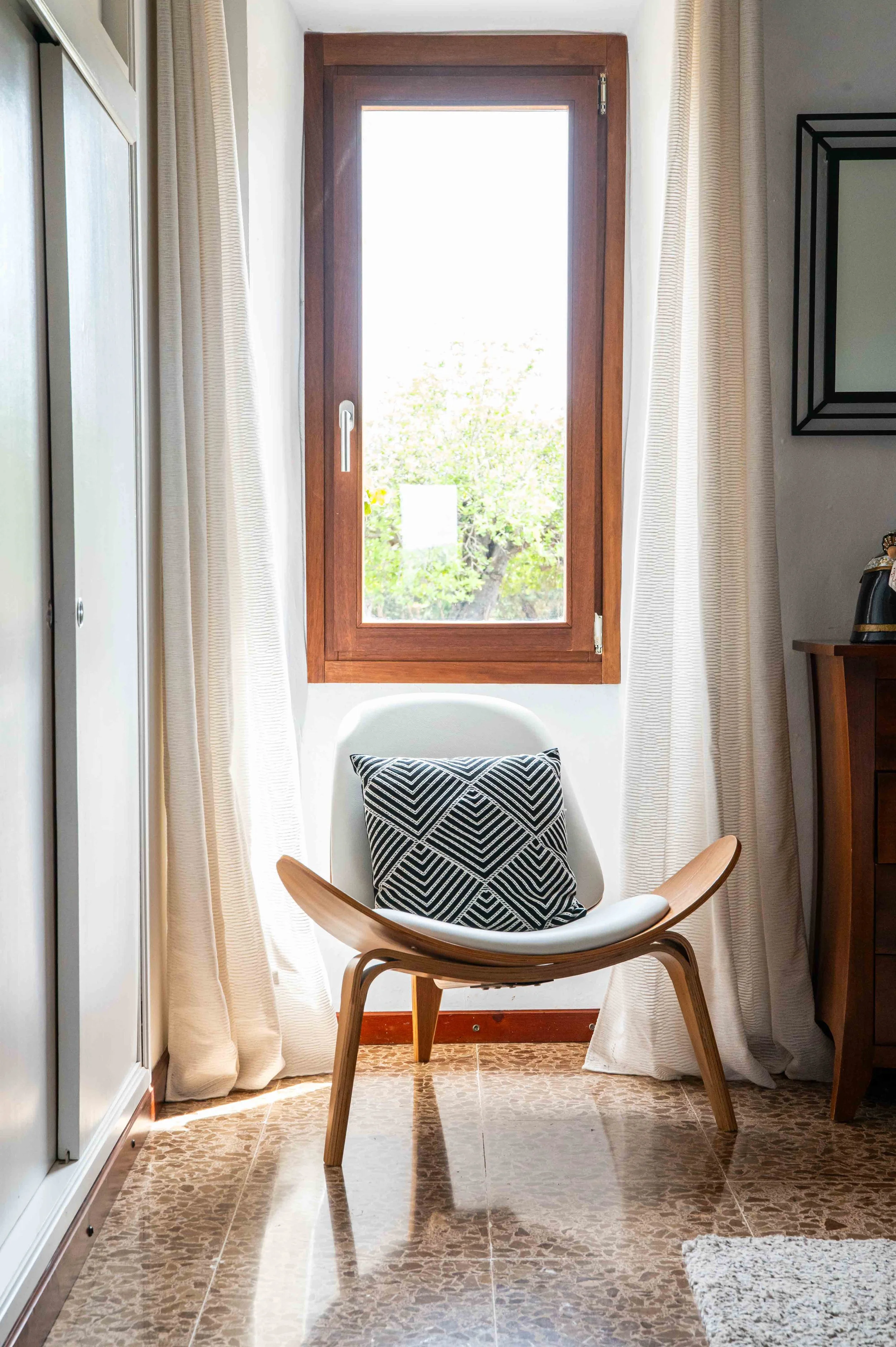 A cozy interior space featuring a white chair with wooden armrests and a black and white geometric patterned pillow. The chair is placed in front of a large window with a wooden frame, flanked by white curtains. The floor is tiled with brown patterne