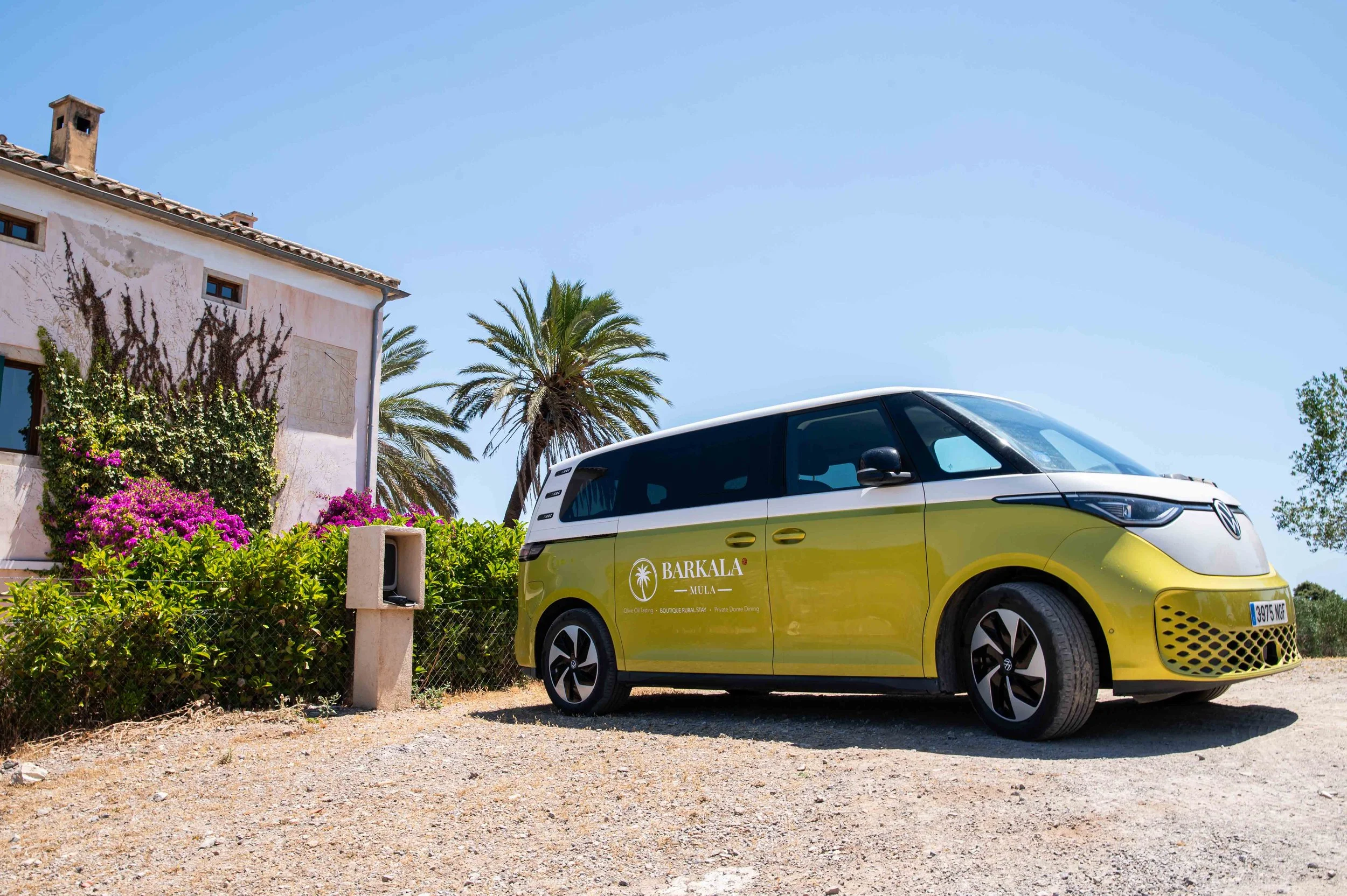 A yellow and white electric vehicle parked outdoors near a building with pink walls and a tiled roof, surrounded by green shrubs and colorful flowers, with palm trees and a clear blue sky in the background.