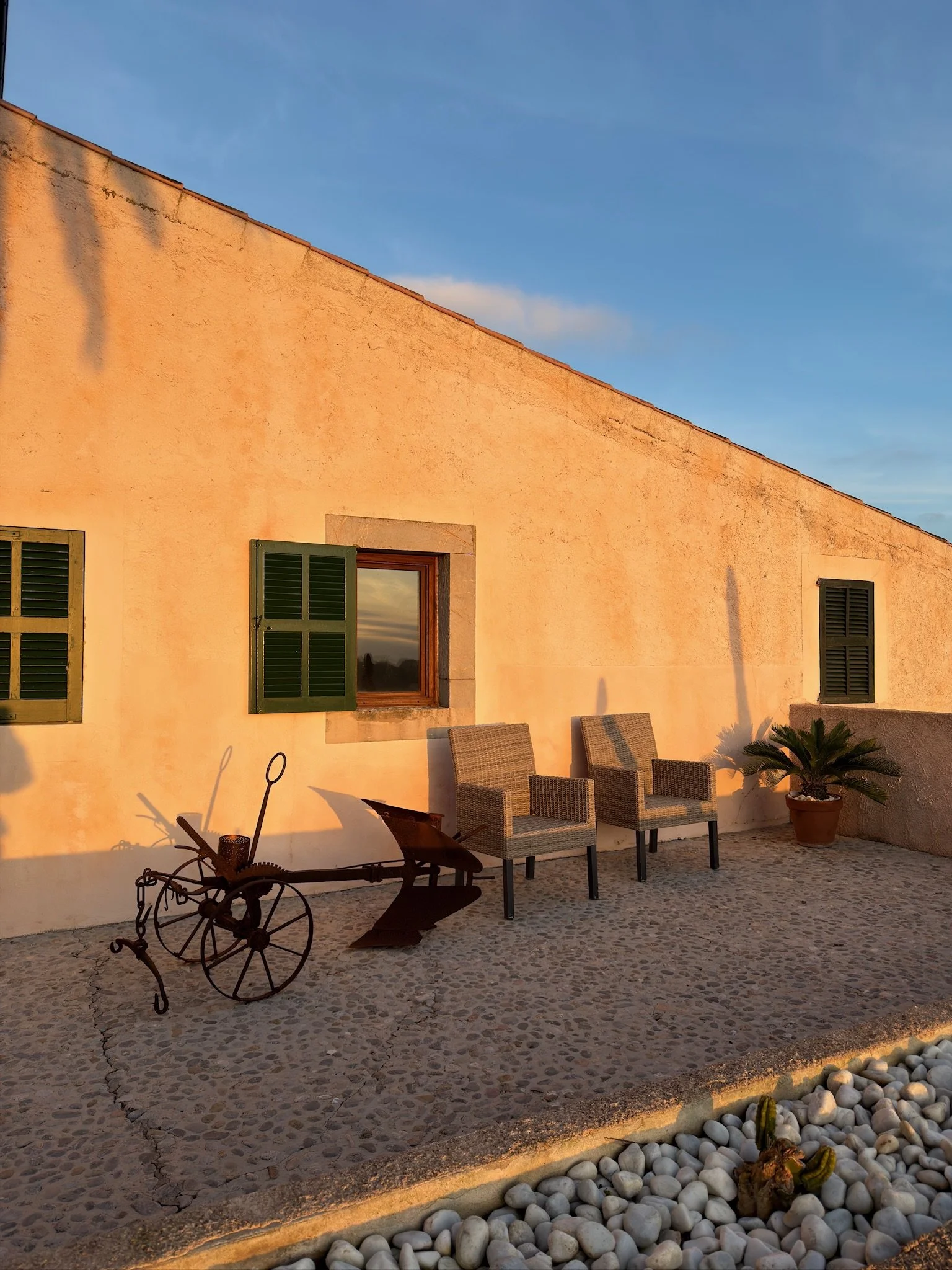 A cozy outdoor patio scene at sunset with a peach-colored stucco house wall, green window shutters, outdoor chairs, decorative plants, a vintage metal bicycle sculpture, and smooth white stones on the ground.