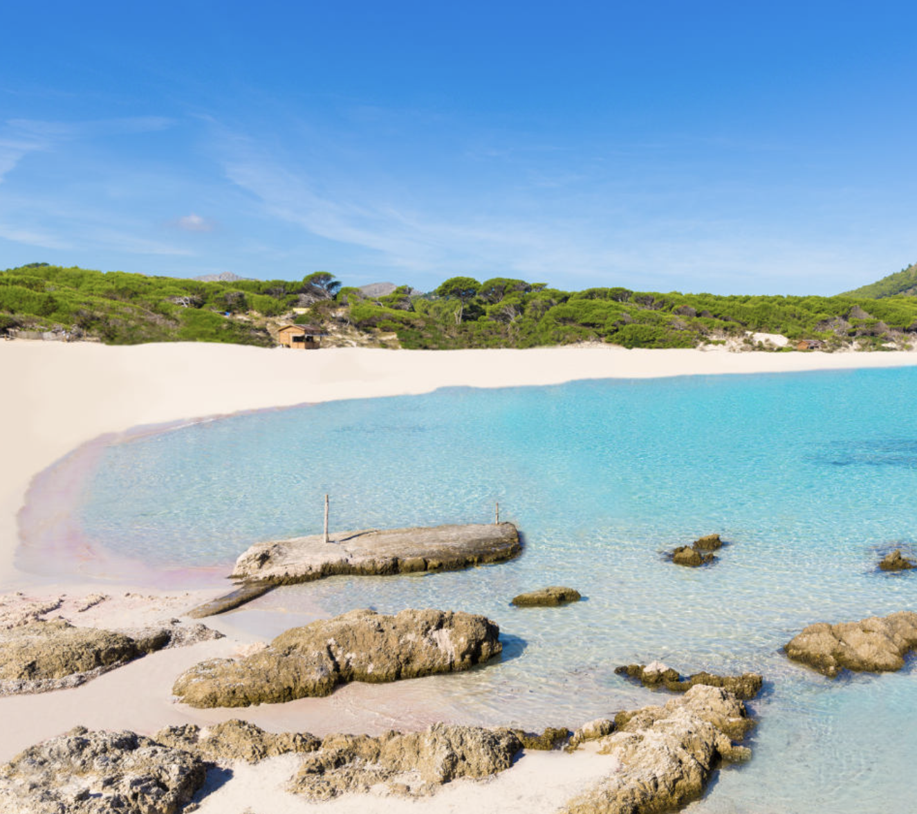 Tropical beach with clear turquoise water, sandy shore, rocks, green trees, houses on hillside, and blue sky with some clouds.