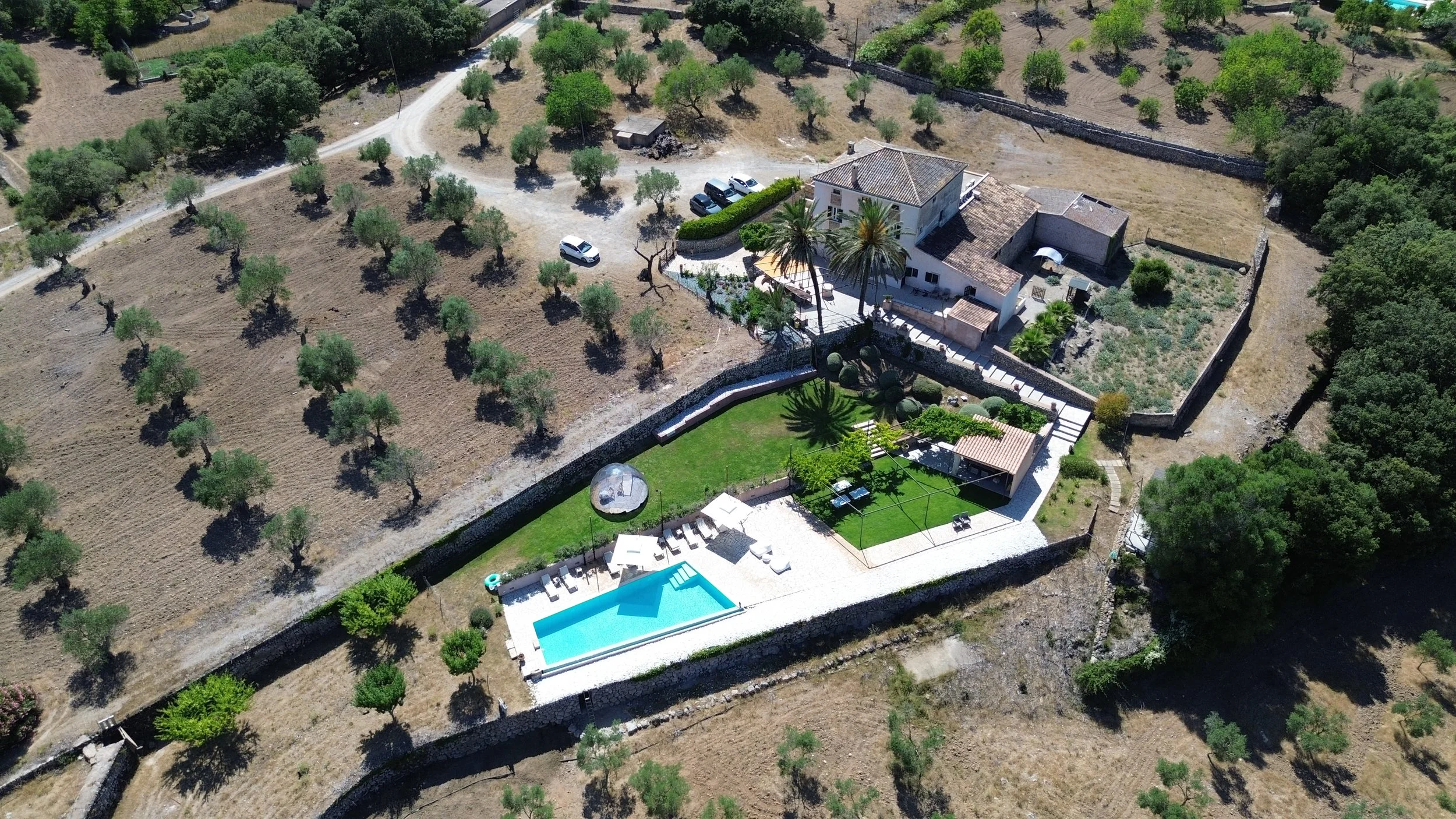 Aerial view of a large house with a swimming pool in the backyard, surrounded by trees, a lawn, and a patio area with outdoor furniture. The house is enclosed by a stone wall, with a driveway and additional parking area nearby. The landscape includes scattered trees and open land.