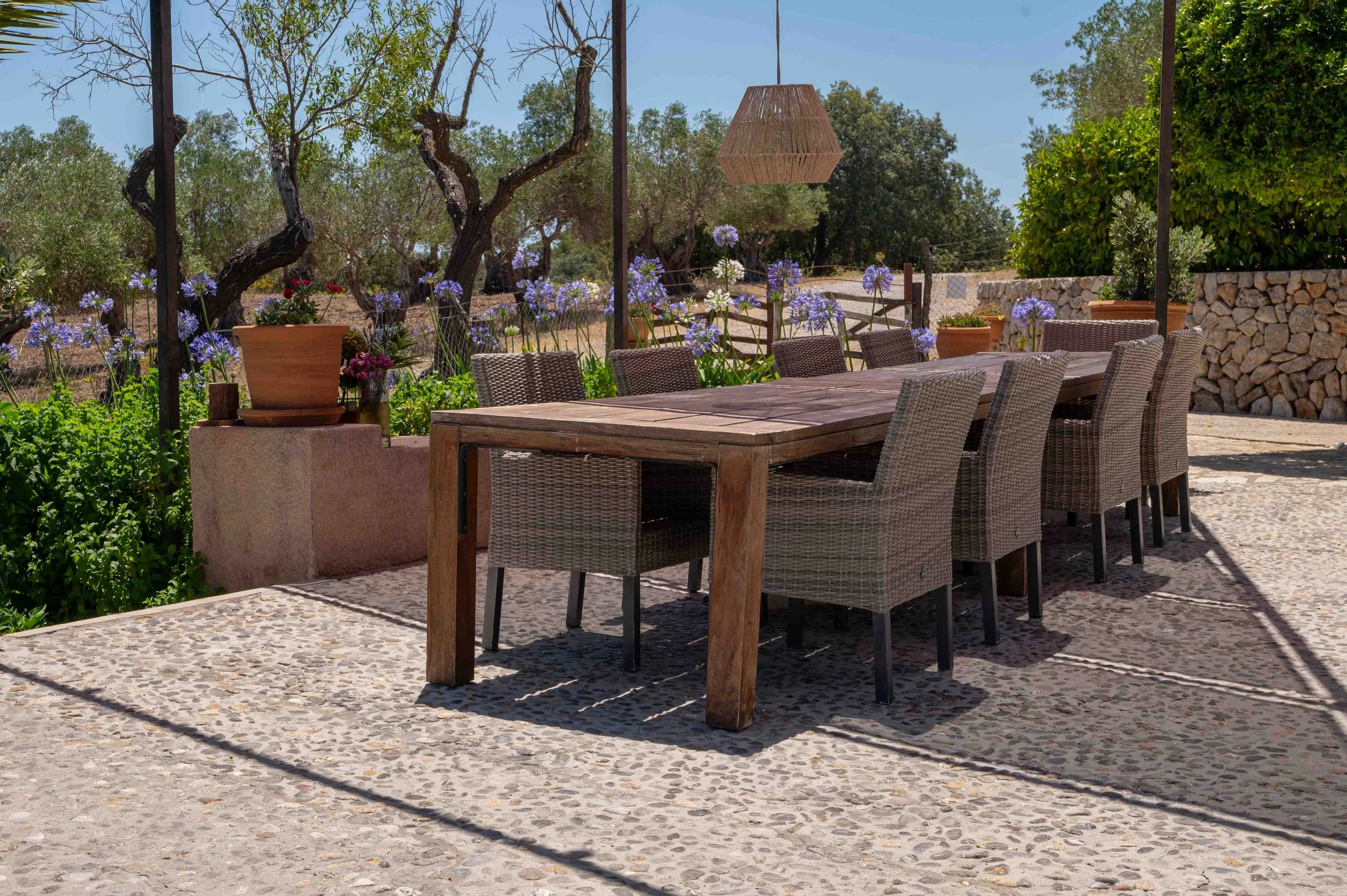 Outdoor dining area with a large wooden table surrounded by woven chairs, potted plants, flowering purple and red plants, trees, a stone wall, and a hanging wooden light fixture, with clear blue sky in the background.