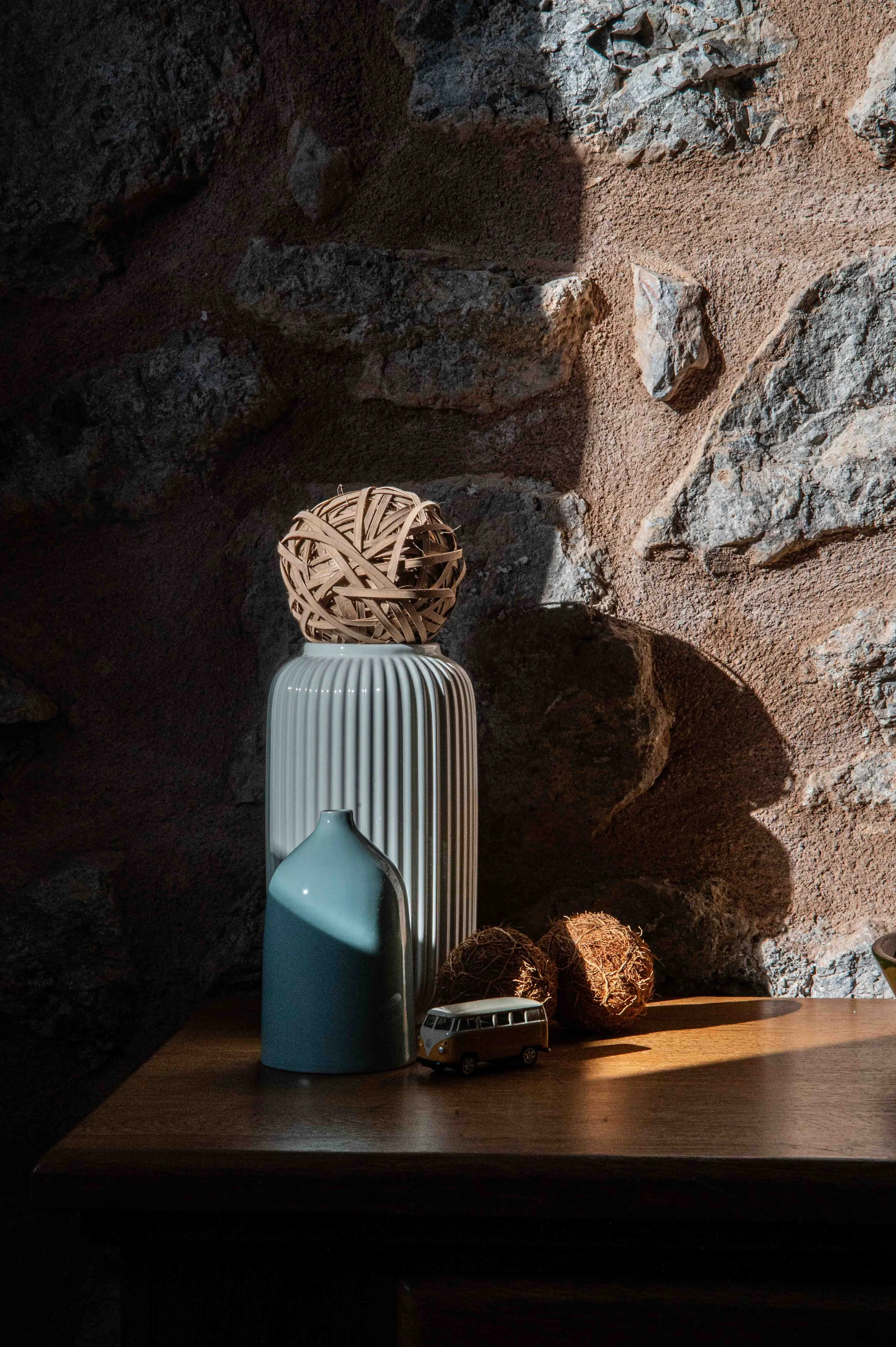 Decorative arrangement on a wooden surface in front of a textured stone wall, featuring a tall ribbed white vase, a small light blue vase, a tan woven decorative object, and a miniature toy bus.