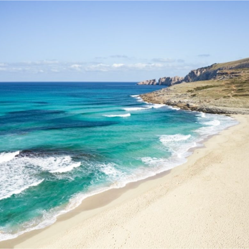 A scenic view of a sandy beach with turquoise ocean waves and rocky cliffs in the background under a partly cloudy sky.