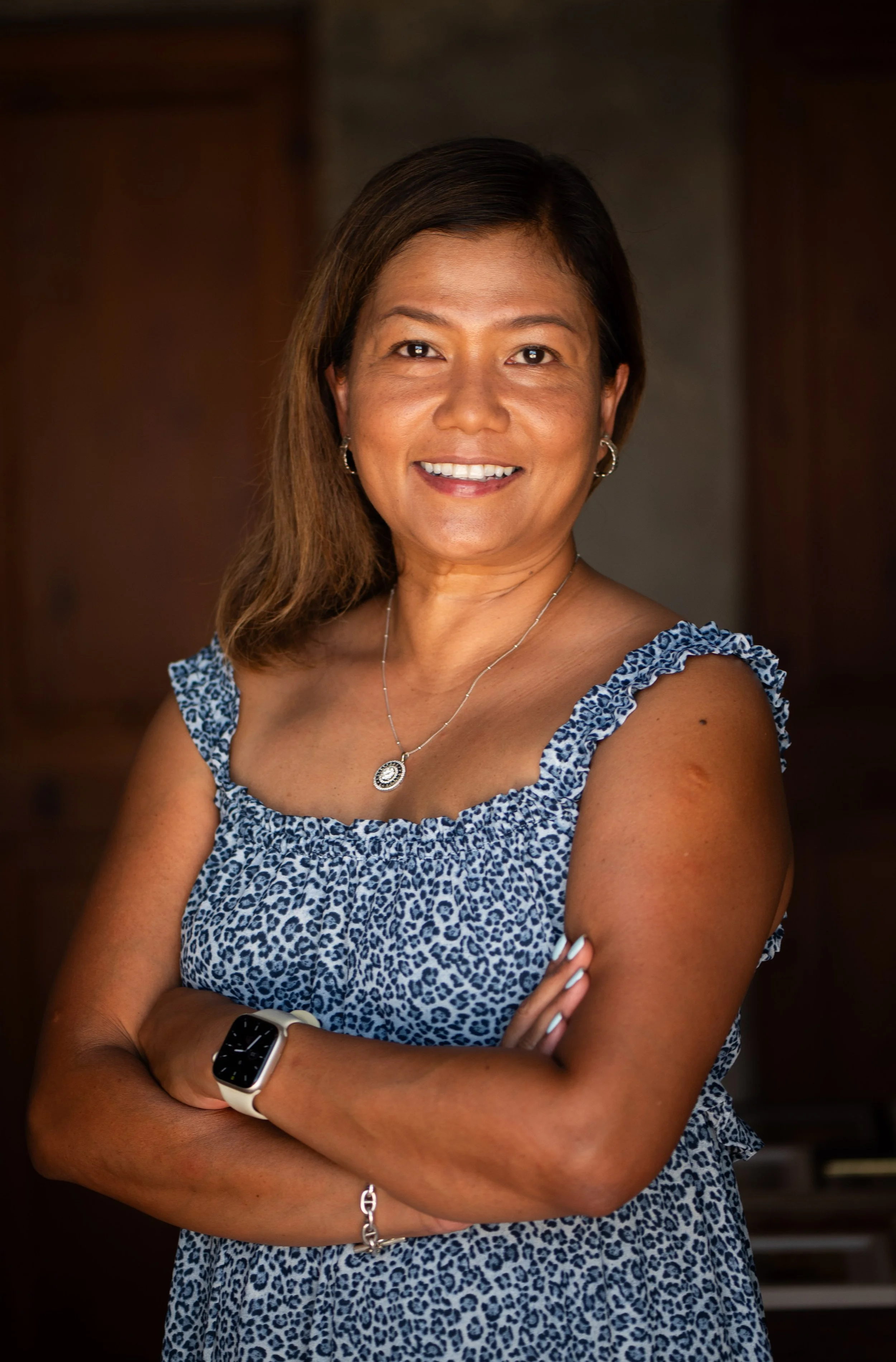A portrait of a woman with shoulder-length brown hair, wearing a blue and white animal print sleeveless dress, a silver necklace with a circular pendant, earrings, an Apple Watch, and a bracelet, smiling with arms crossed against a dark indoor background.