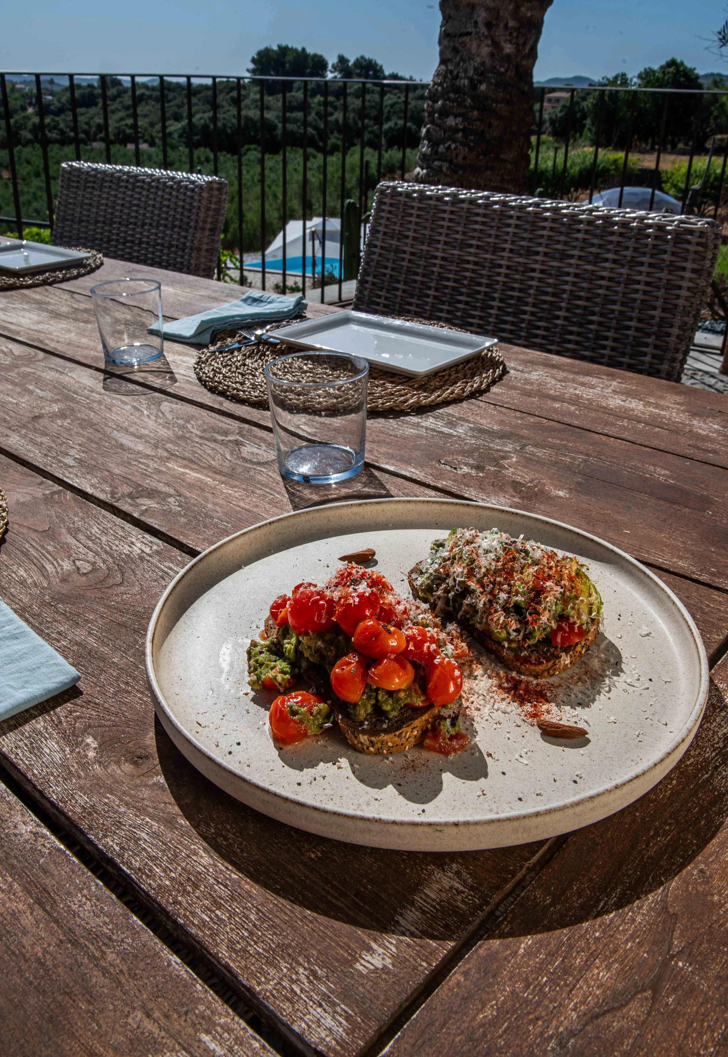 A rustic outdoor dining table set with decorative placemats, dishes, glasses, and napkins. The background features a scenic view of trees, a swimming pool, and rolling hills under a clear blue sky.