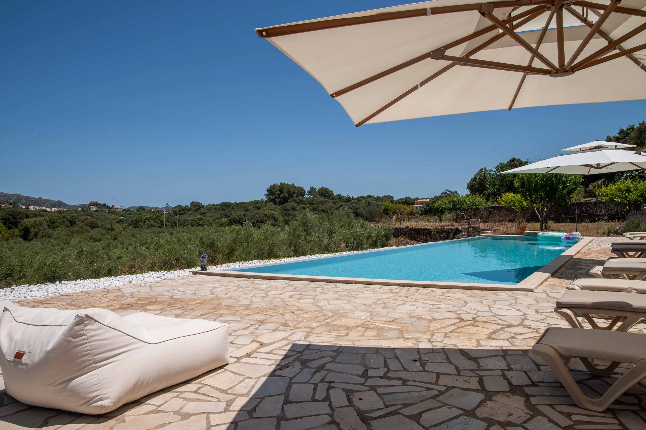 An outdoor swimming pool surrounded by lounge chairs and umbrellas, set against a scenic landscape of hills and greenery under a clear blue sky.