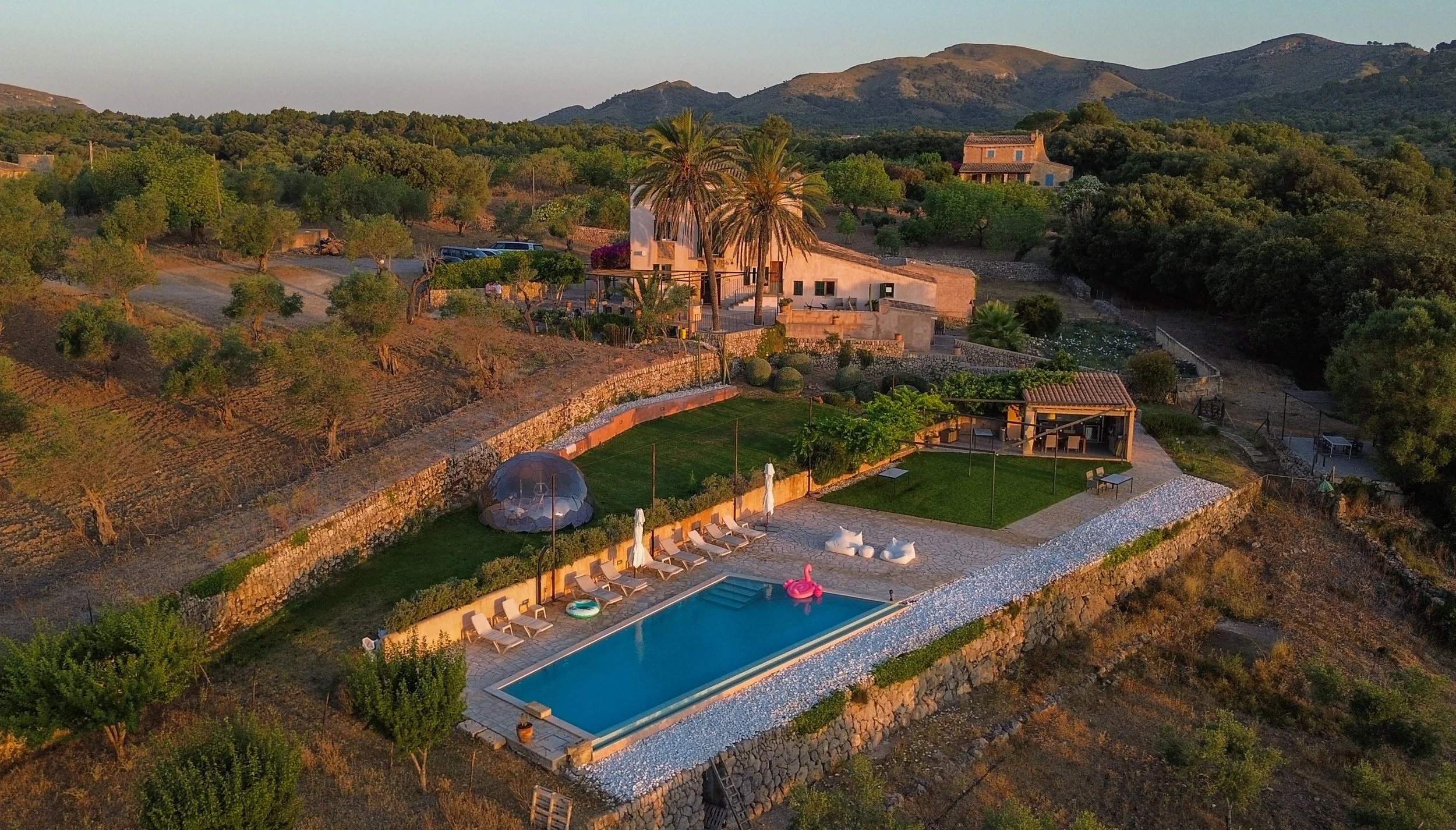 Aerial view of a hillside estate with a swimming pool, lounge chairs, a shaded patio, lush vegetation, trees, and hills in the background during sunset.