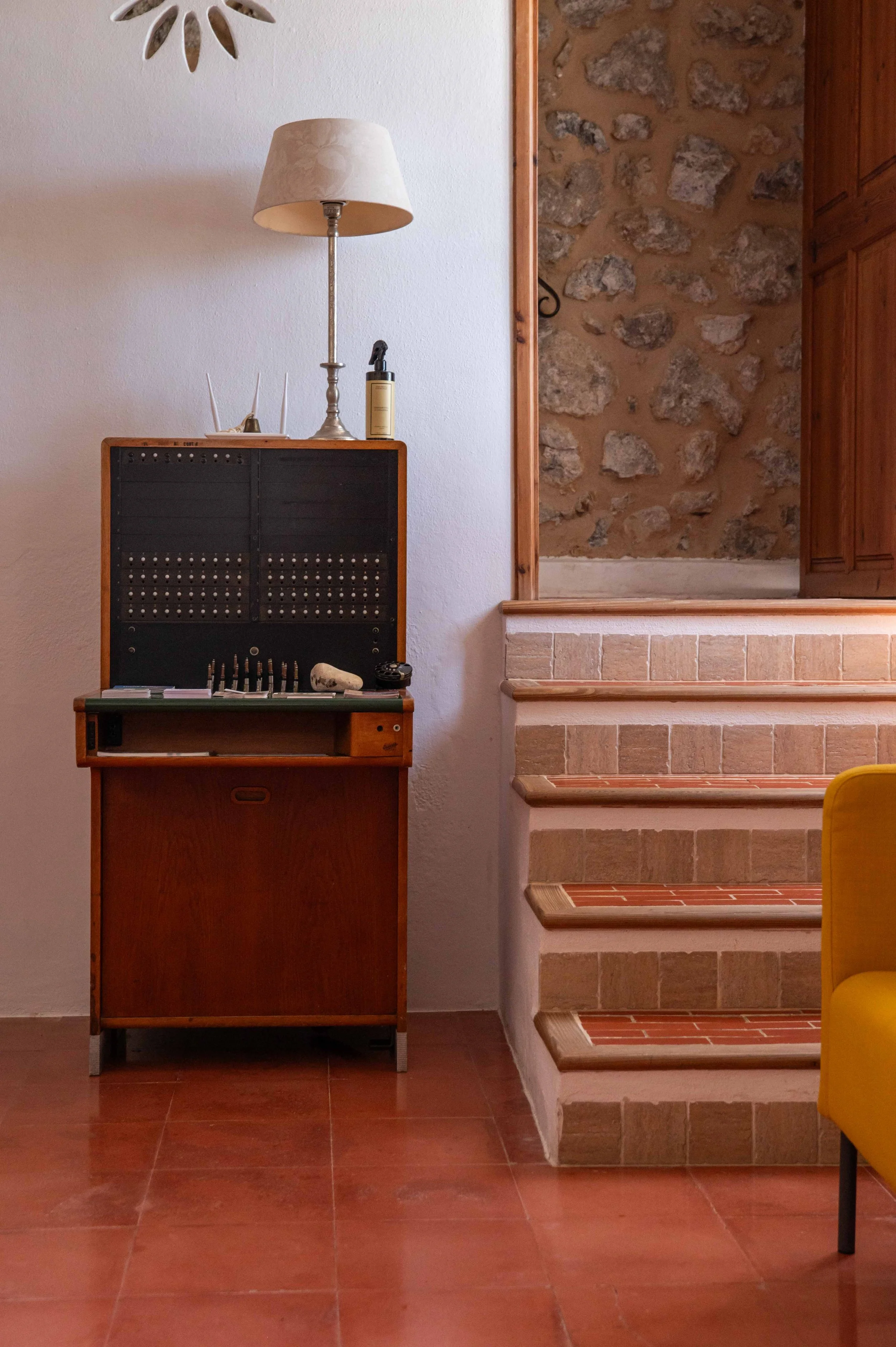A wooden cabinet with a black panel on the front, topped with a table lamp and a bottle. To the right, there are stairs with brick and wood steps leading to a upper level, and a stone wall visible through an opening. Part of a yellow chair is seen on