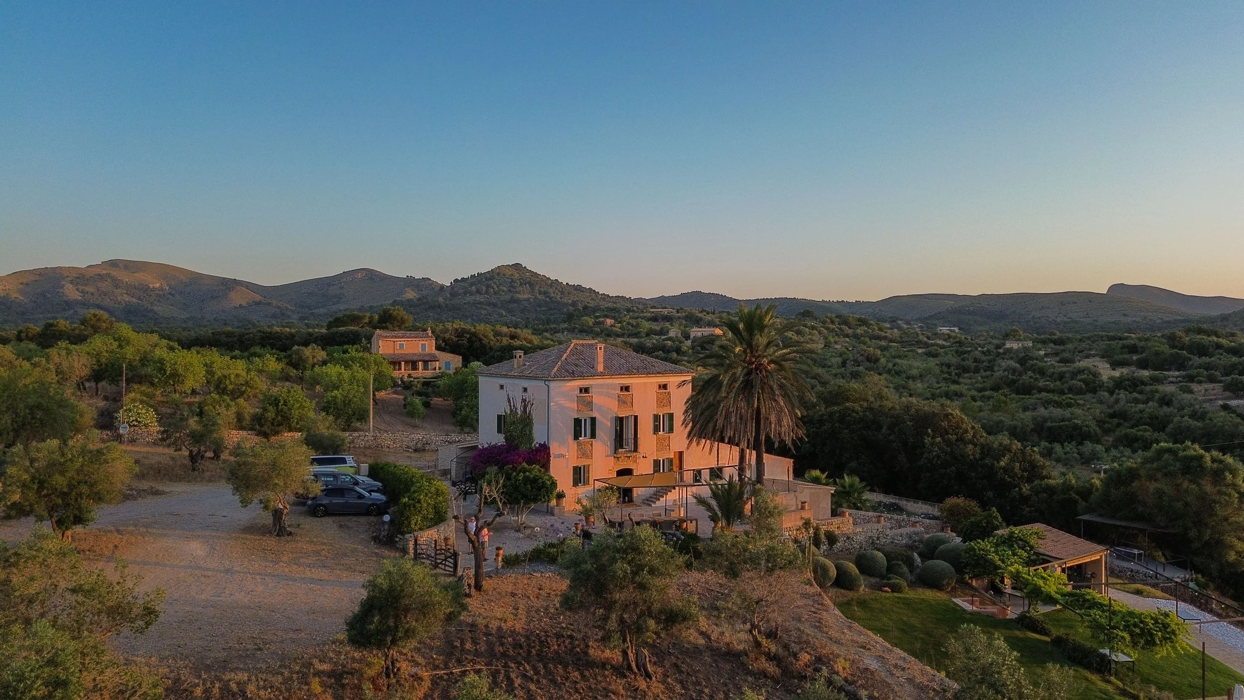 A scenic view of a hillside with houses, trees, and mountains in the background at sunset.