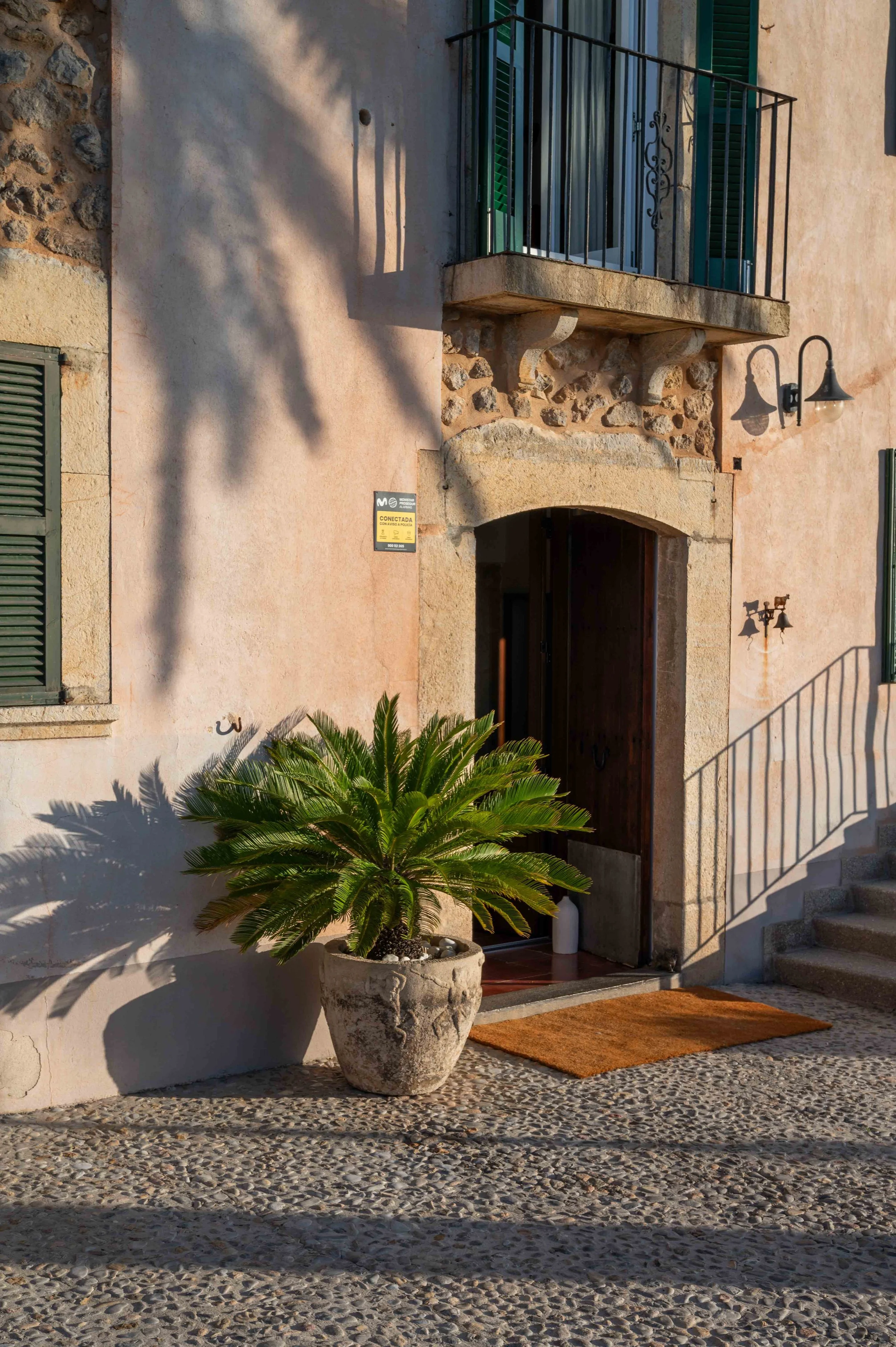 A building house with a stone and stucco facade, green shutters on the windows, a small balcony with a black metal railing, two black outdoor wall lamps, a large potted palm plant in front, a doormat, and a staircase to the right.