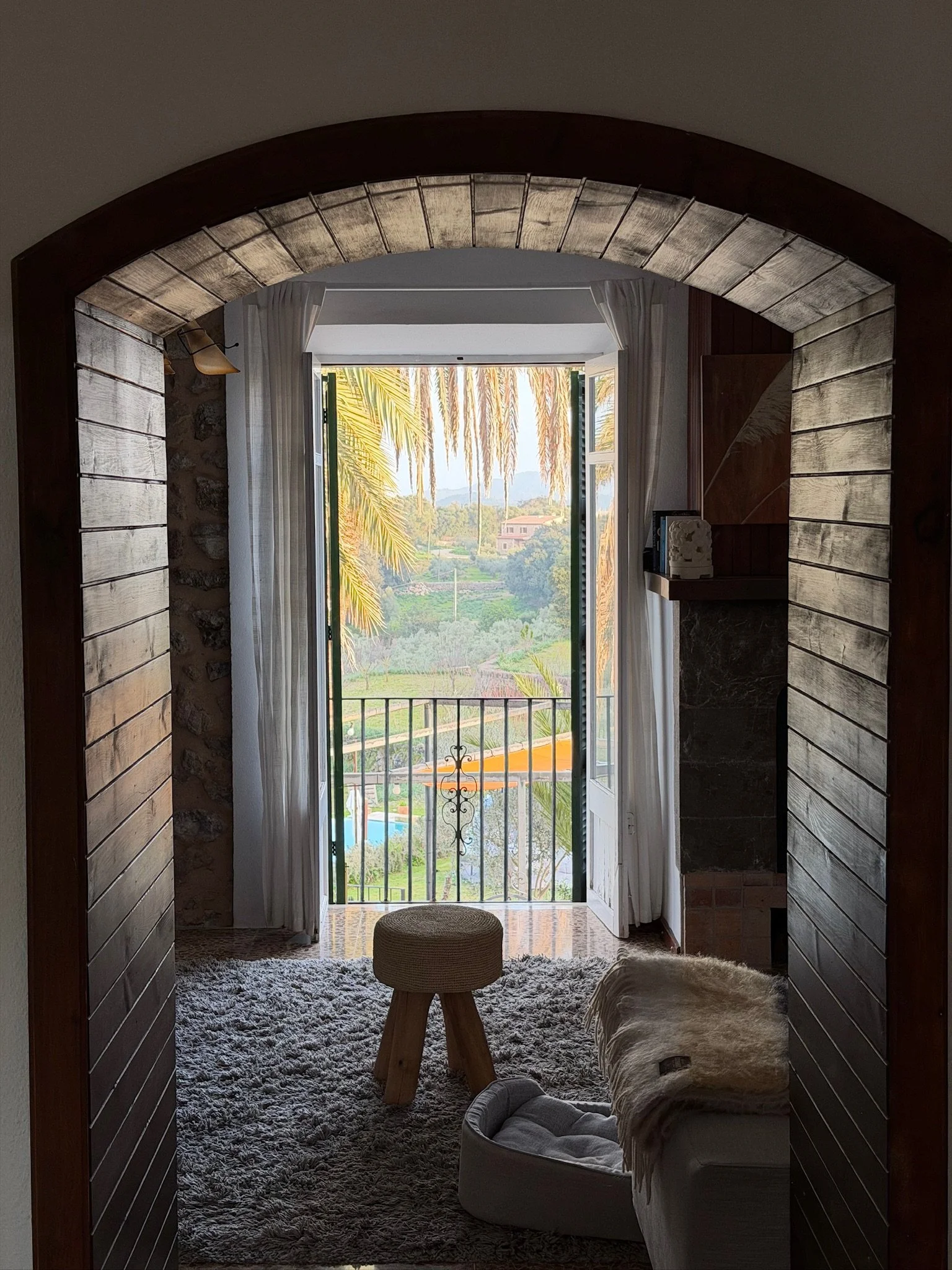 View from inside a room looking out through an open glass door with white curtains, overlooking a balcony with a view of palm trees and greenery in the distance, during daytime.