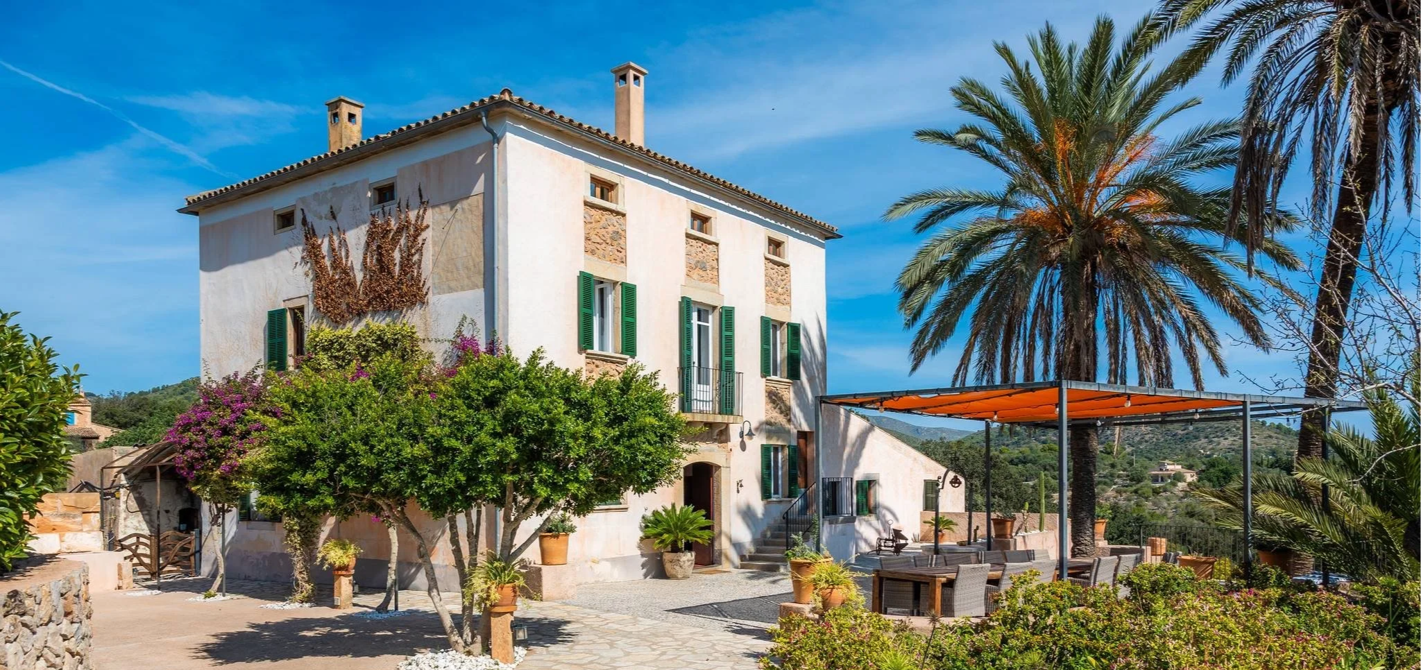 A white Mediterranean-style house with green shutters, surrounded by lush greenery and tall palm trees, under a clear blue sky.