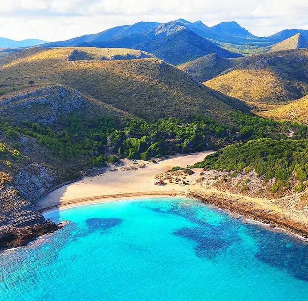 A secluded beach with clear blue water, a sandy shore, surrounded by green trees and dry hills with mountains in the background.