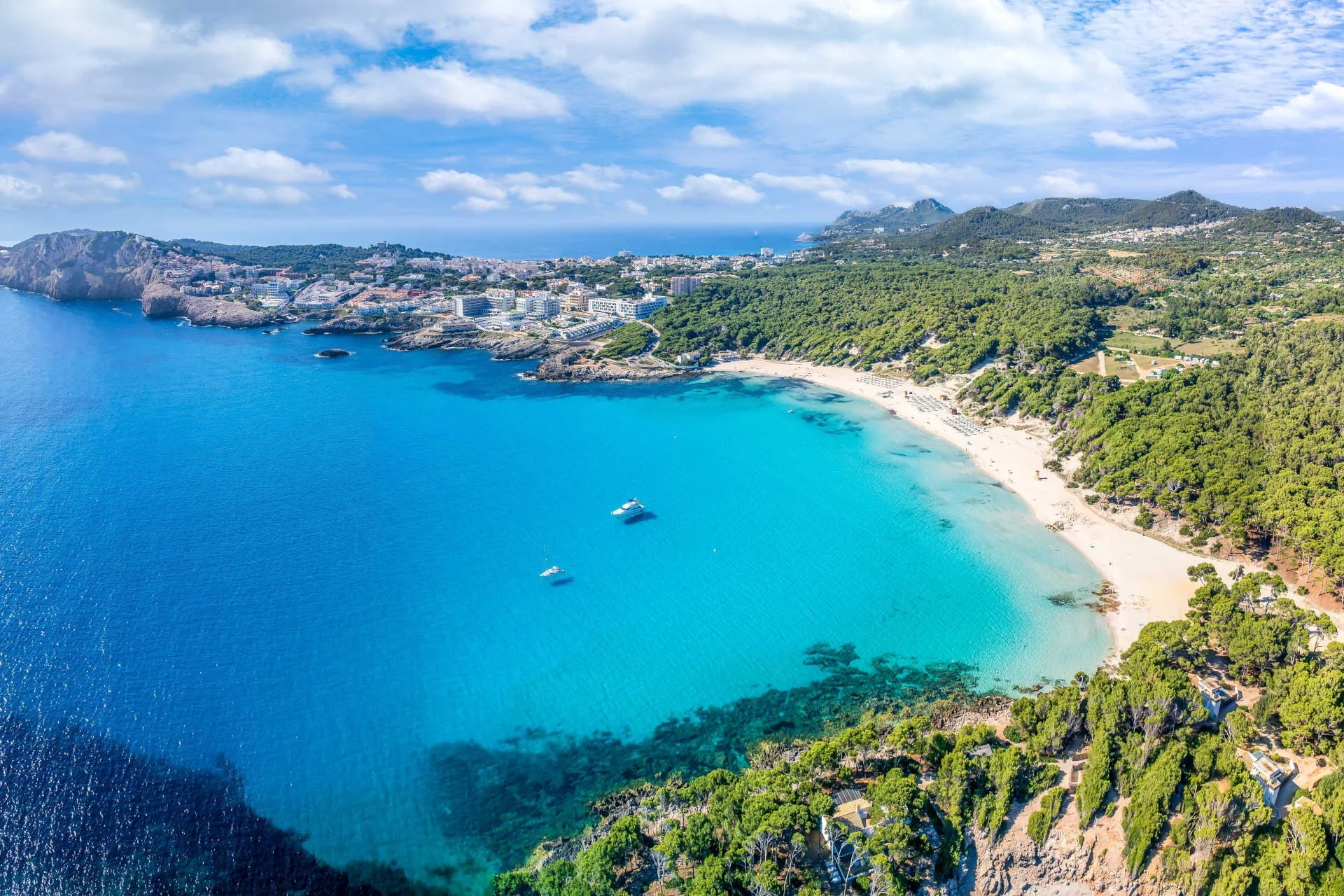 Aerial view of a scenic coastline with turquoise waters, sandy beach, lush green hills, and a coastal town under a partly cloudy sky.