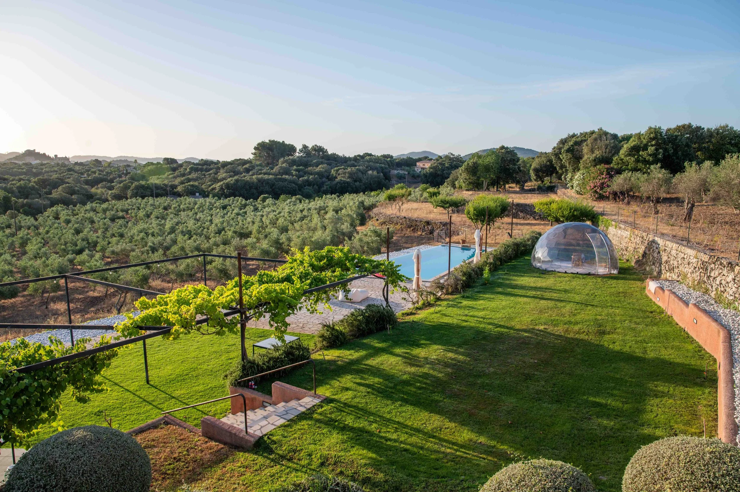 A scenic backyard with a green lawn, a small pool, a transparent dome structure, and planting beds, overlooking a hilly landscape with trees and distant mountains.