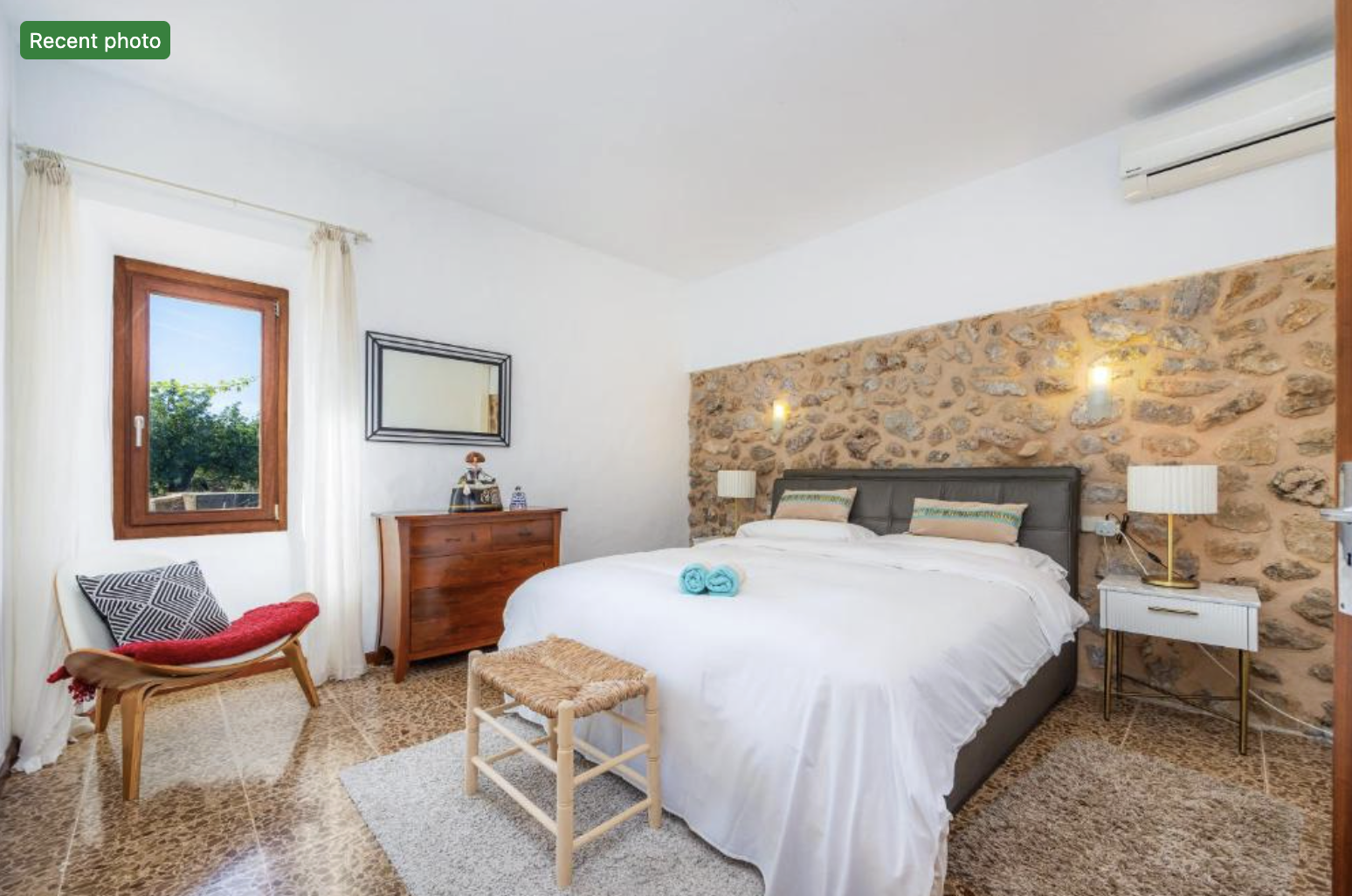 A bedroom with a large bed, stone accent wall, wooden furniture, a window with white curtains, and various decorative items.