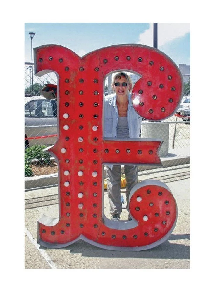 A woman stands behind a large vintage red letter "E" with embedded bulbs, smiling while looking through the letter.