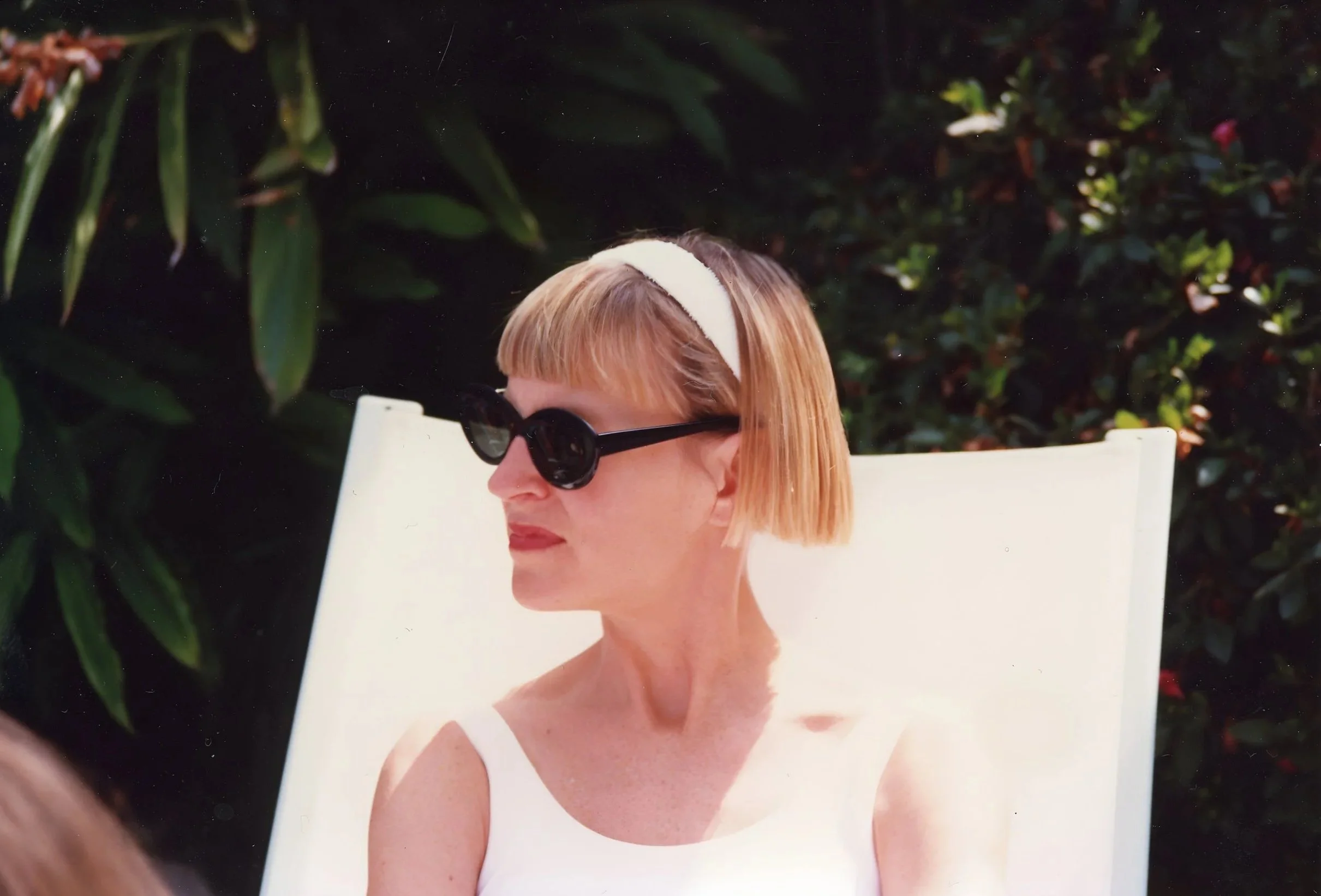 A woman with short blonde hair and black sunglasses sits outdoors against a backdrop of green foliage, wearing a white sleeveless top.