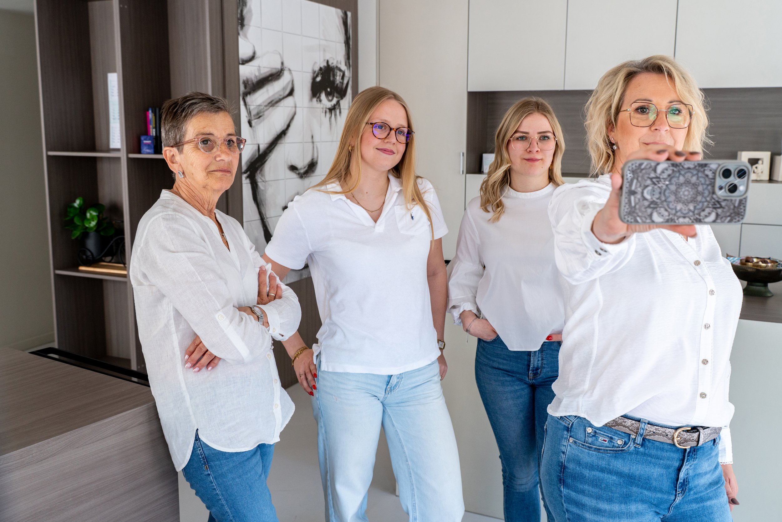 Vijf vrouwen poseren voor een foto in een moderne keuken, waarbij de vrouw aan de rechterkant een selfie neemt.