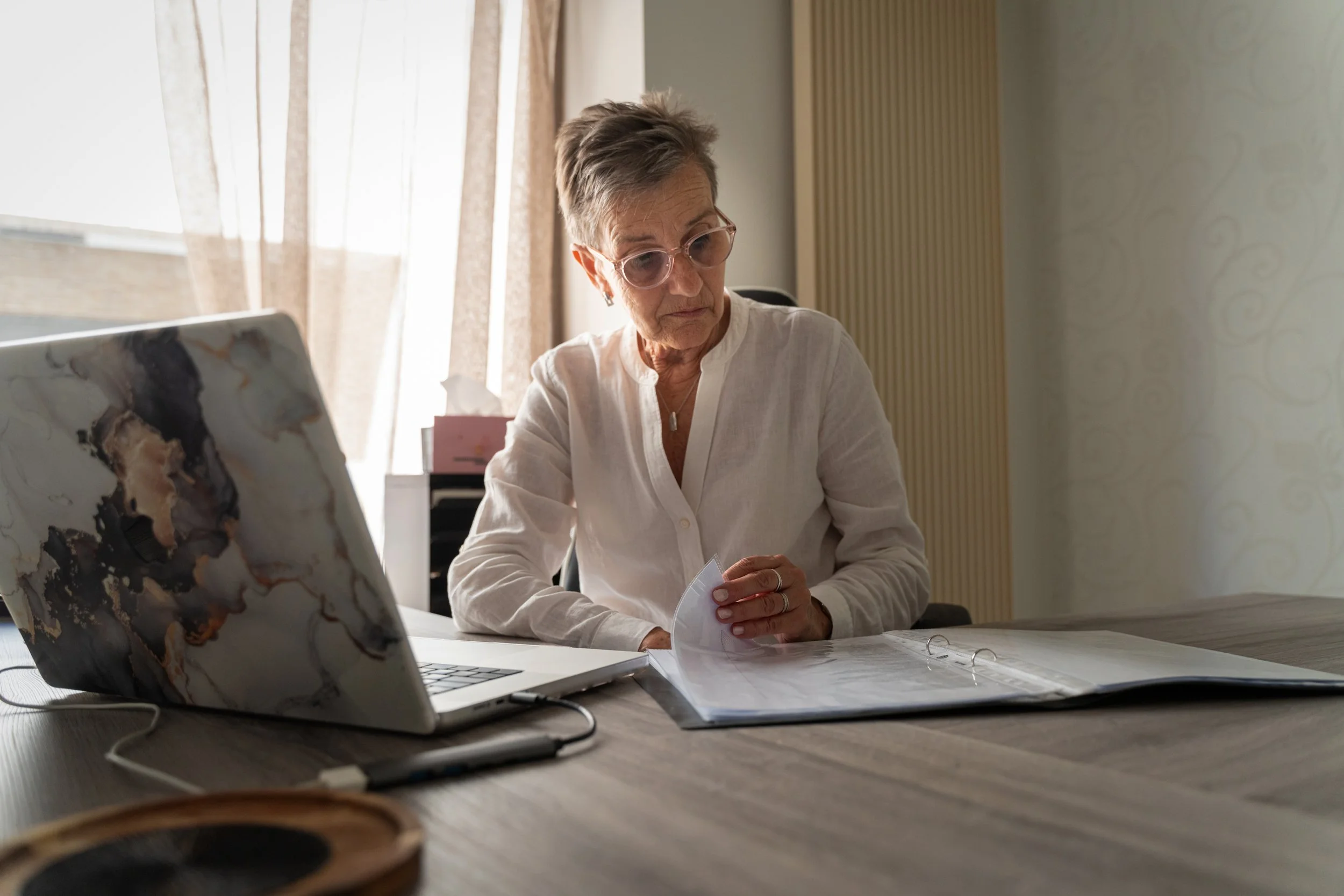 Een oudere vrouw met kort grijs haar, draagt een witte blouse en bril, zit aan een bureautafel en kijkt naar documenten, met een laptop en een tablet op de tafel.